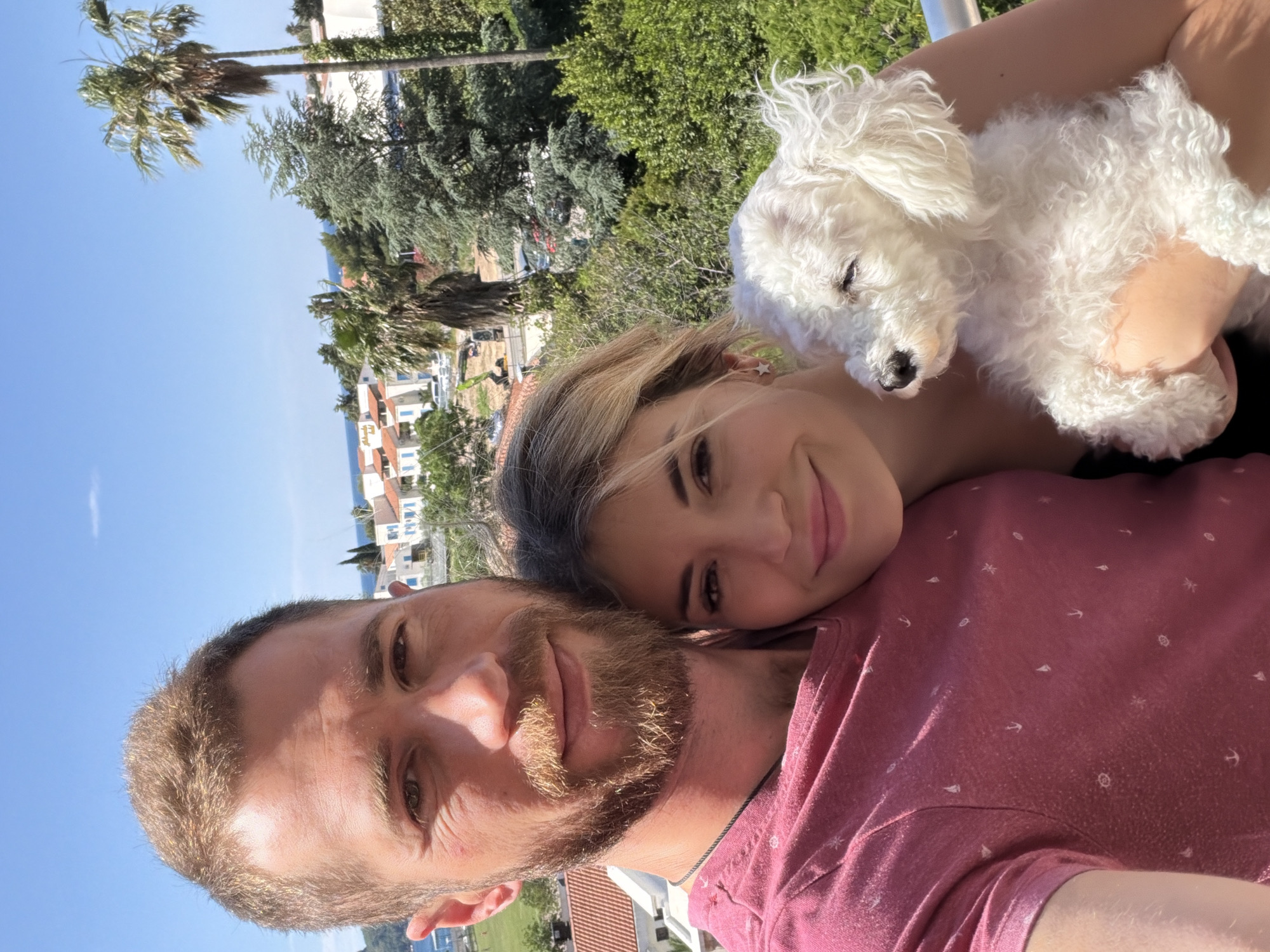 A selfie photograph depicts a man and a woman, with a small white dog. The man, in the foreground, has short hair and a beard, wearing a red shirt with small white designs. The woman is behind the man and slightly to the side, with blonde hair and a pink complexion. A small white dog is resting in the woman’s arms. The background includes vegetation such as trees, palm trees, and buildings. The sky is blue. The scene is likely located in Budva, Montenegro, given the architectural style of the buildings and the surrounding landscape.