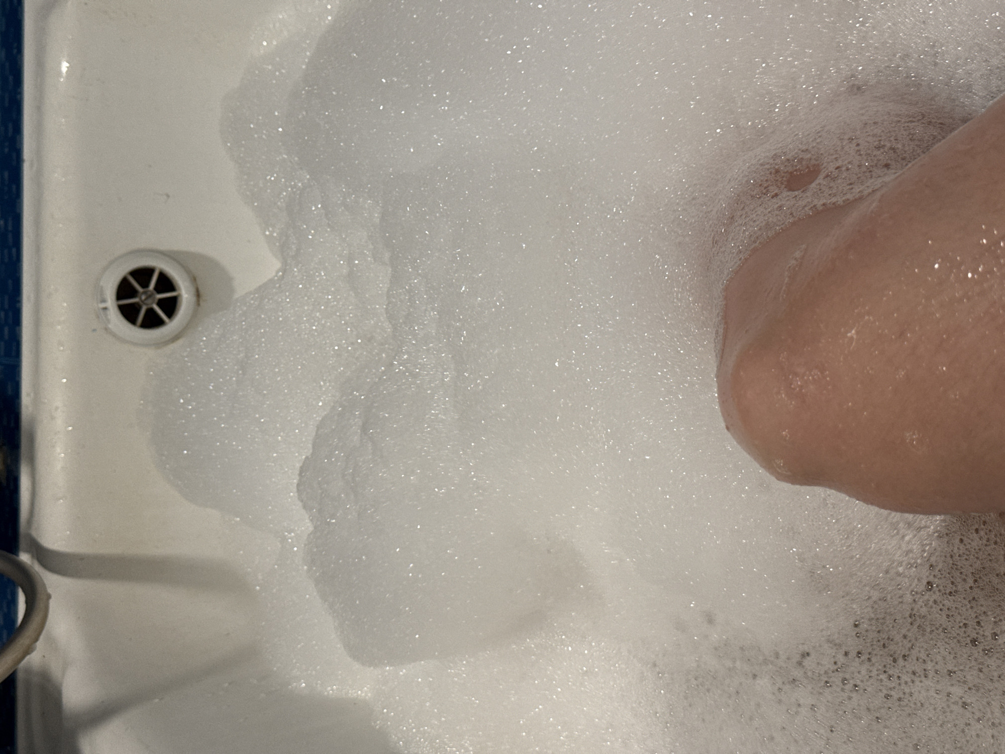 The image shows a white bathtub filled with white bubbles, with the drain visible on the left side. A person's leg is partially visible on the right side of the frame, submerged in the bubbles. The photo appears to be taken from a high angle, looking down into the tub. There is no information available about the location being in the city of Chaikovsky, Russia, so this cannot be determined from the image.