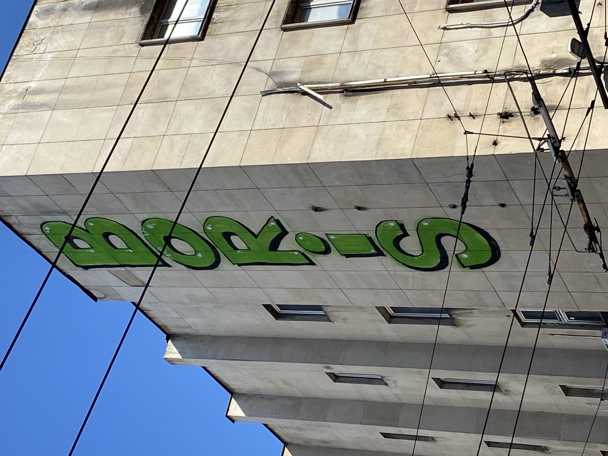The image depicts a section of a tall, concrete building, most likely located in Sofia, Bulgaria, given the Cyrillic-style graffiti. The building's exterior is characterized by a grid of square panels, some of which contain small, rectangular windows. Prominently displayed on the building's facade is a large, green graffiti tag reading "BORIS" in stylized block letters with black outlines. Power lines and electrical equipment are visible in the foreground and to the right, obscuring part of the building. The sky is clear and blue. The overall perspective is low, looking up at the building, which seems to overhang the viewer's position.