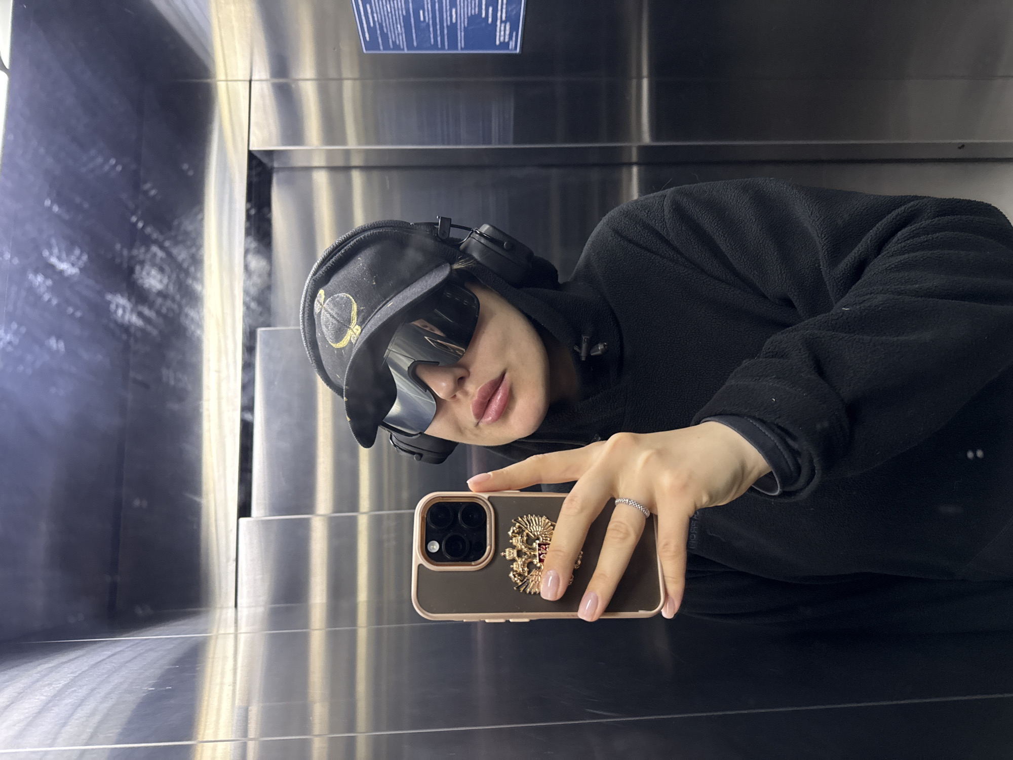 A woman takes a selfie in what appears to be the interior of an elevator. The walls of the elevator have a metallic, brushed silver appearance. The woman is wearing a black hooded jacket, a black baseball cap with a yellow emblem, sunglasses, and headphones. Her left hand holds a smartphone with a case displaying a golden emblem featuring a double-headed eagle design. The woman is wearing a ring on her ring finger. A blue sign is visible near the top of the elevator. This image was likely taken in Krasnoyarsk, Russia, based on the emblem on the smartphone case.