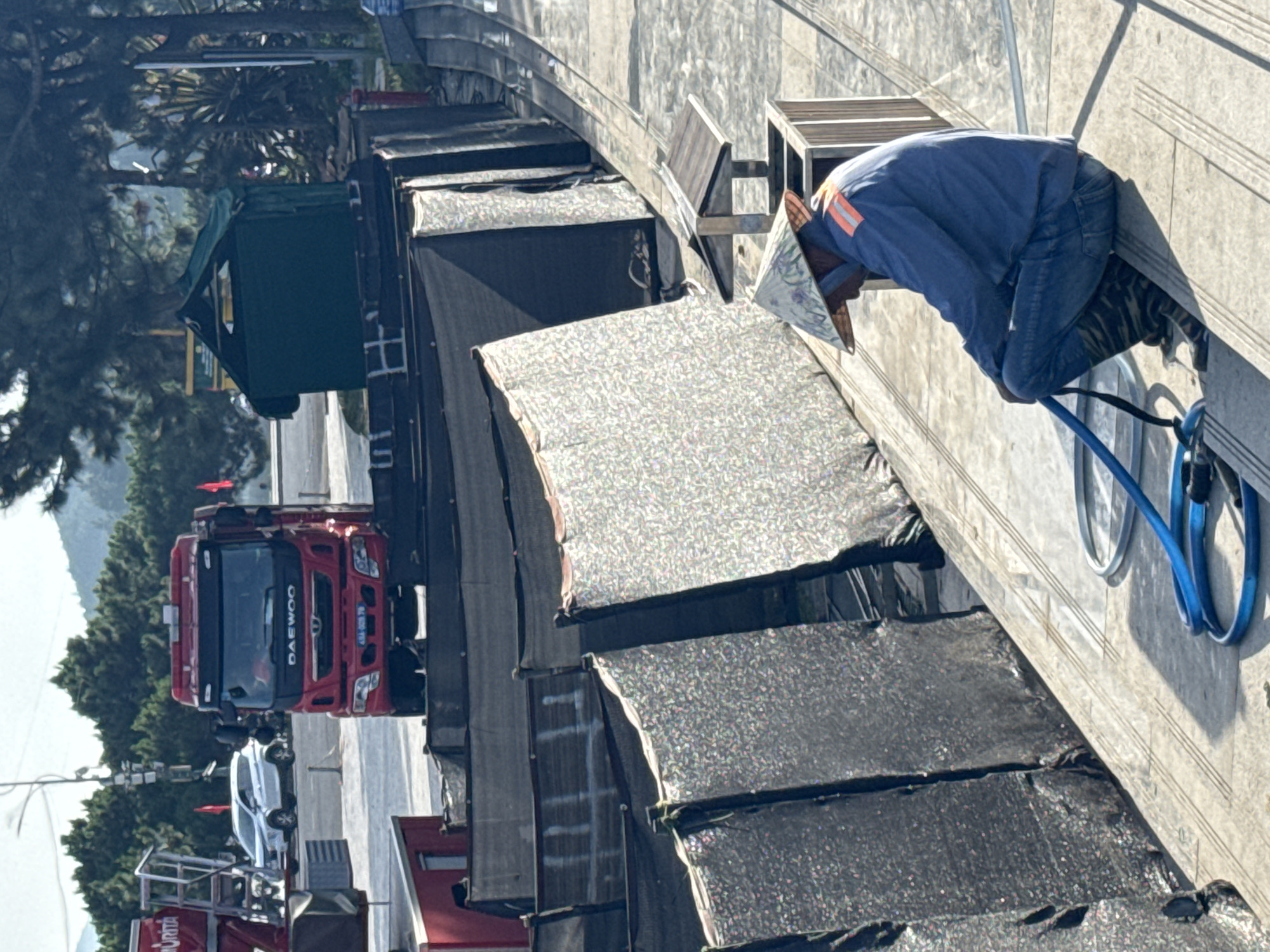 The image depicts an outdoor scene, possibly a construction site or a work area, with a man in the foreground. He is wearing a conical hat, a blue jacket with orange reflective stripes on the shoulders, blue jeans, and patterned boots. The man is crouching and holding a blue hose. Behind him, there are rows of black mesh fencing with some sections covered in a sparkly, reflective material. In the middle ground, a red Daewoo truck is parked, with a white SUV and a green tent-like structure visible further behind. The background features lush green trees and a slightly hazy sky, suggesting a location with natural surroundings. The setting appears to be a relatively open area within a city, given the presence of vehicles and infrastructure. Without further context or identifying landmarks, pinpointing the exact location within Da Lat, Vietnam is not possible solely from the visual information.