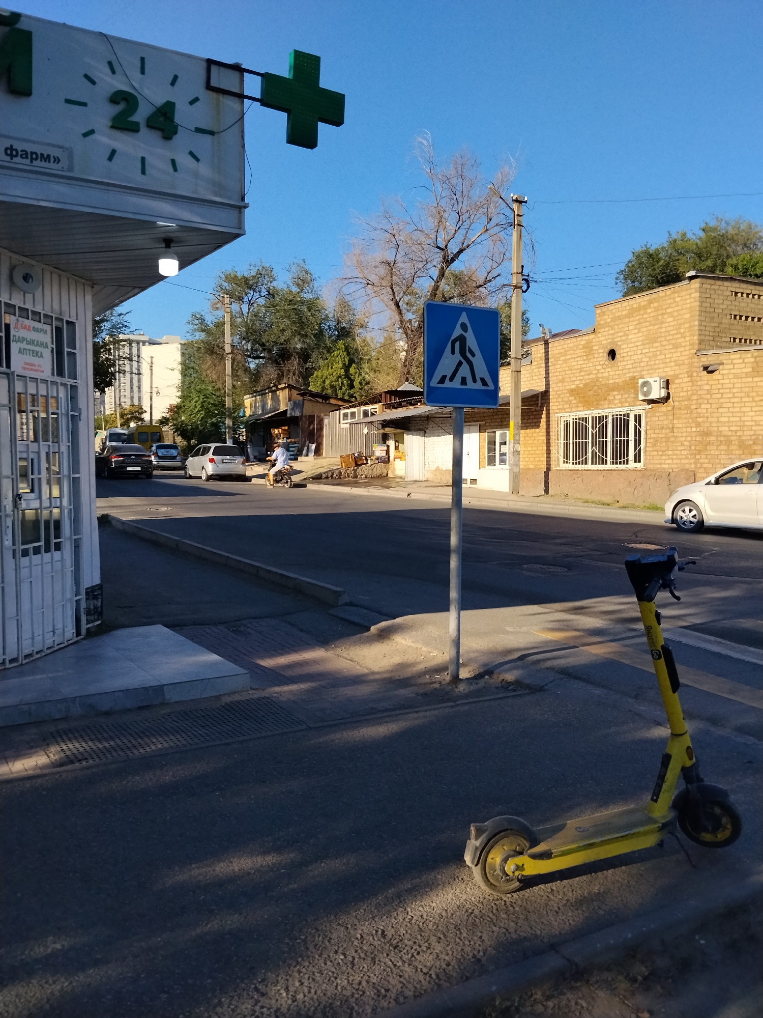 The image depicts a street scene in Bishkek City, Kyrgyzstan, with a focus on a yellow electric scooter parked on a sidewalk. In the background, a brick building with barred windows is visible, alongside smaller structures and trees. Across the street, cars are parked and driving, and a person is seen riding a motorbike. A pedestrian crossing sign is prominently displayed on a pole. A clock indicating "24" and a green cross sign, likely for a pharmacy, are mounted on a building to the left. The sky is clear and blue, suggesting a sunny day. The overall environment appears to be an urban or semi-urban area.