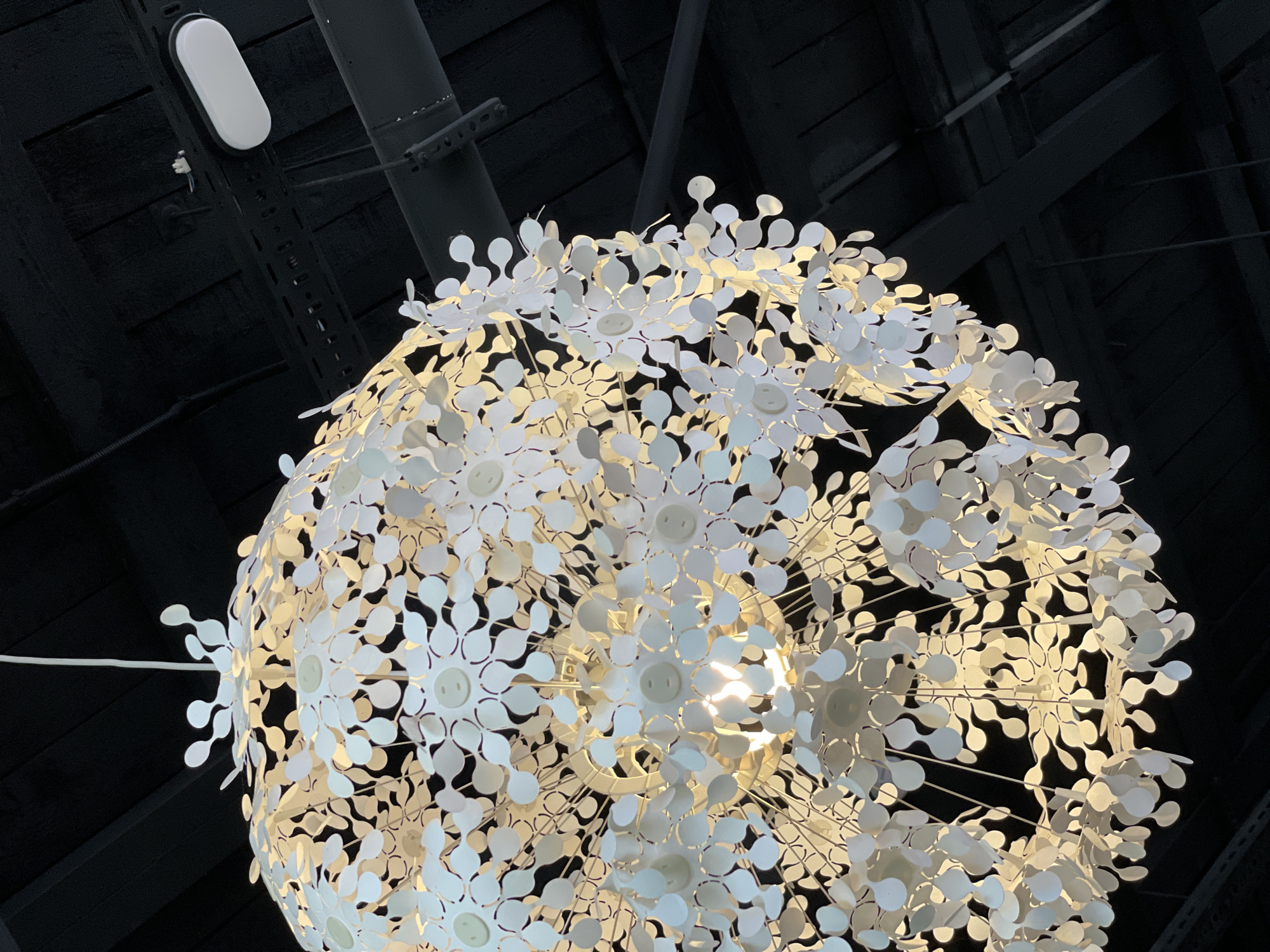 An elaborate white floral chandelier hangs from a dark wooden ceiling with exposed beams. The chandelier is spherical, composed of numerous small, petal-shaped white elements, each with a small circular detail, possibly a button, at its center. A bright light source is visible within the chandelier, illuminating the petals and casting a warm glow. A white cord suspends the fixture. In the upper right portion of the frame, a white oval light fixture is attached to a metal beam that runs along the ceiling. The background is a dark, textured wooden ceiling. There are no people or animals present in the image. This scene is not specific to Krasnoyarsk, Russia, as no identifying landmarks or context are provided.