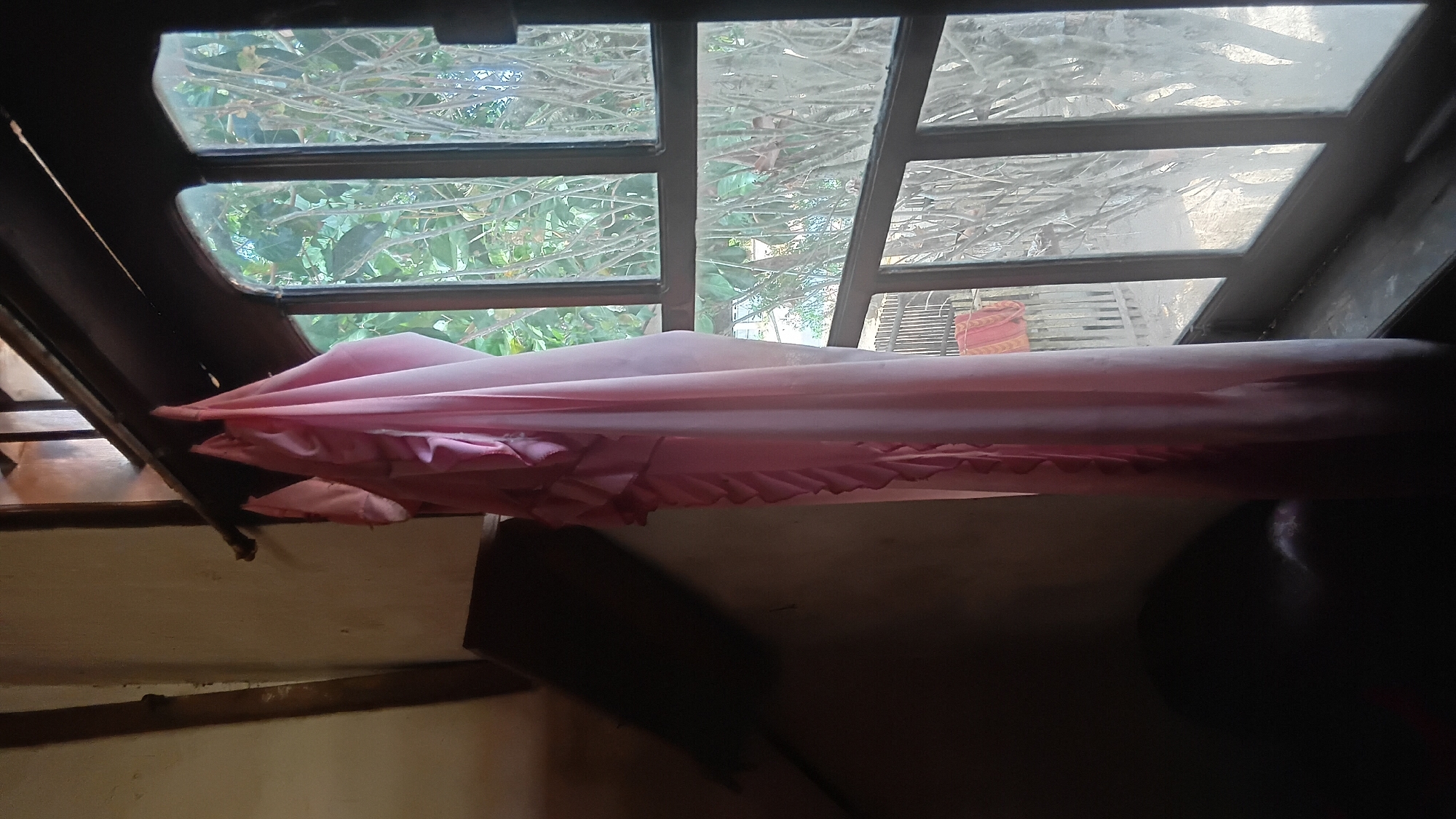 A pink fabric is draped over a rod inside a room, partially obscuring a window. The window is divided into multiple panes and looks out onto trees and a fence. To the left of the fabric, a dark object, possibly furniture, is visible. The wall of the room is a neutral color and appears to have some rough texture. The scene is dimly lit, with light filtering in through the window.