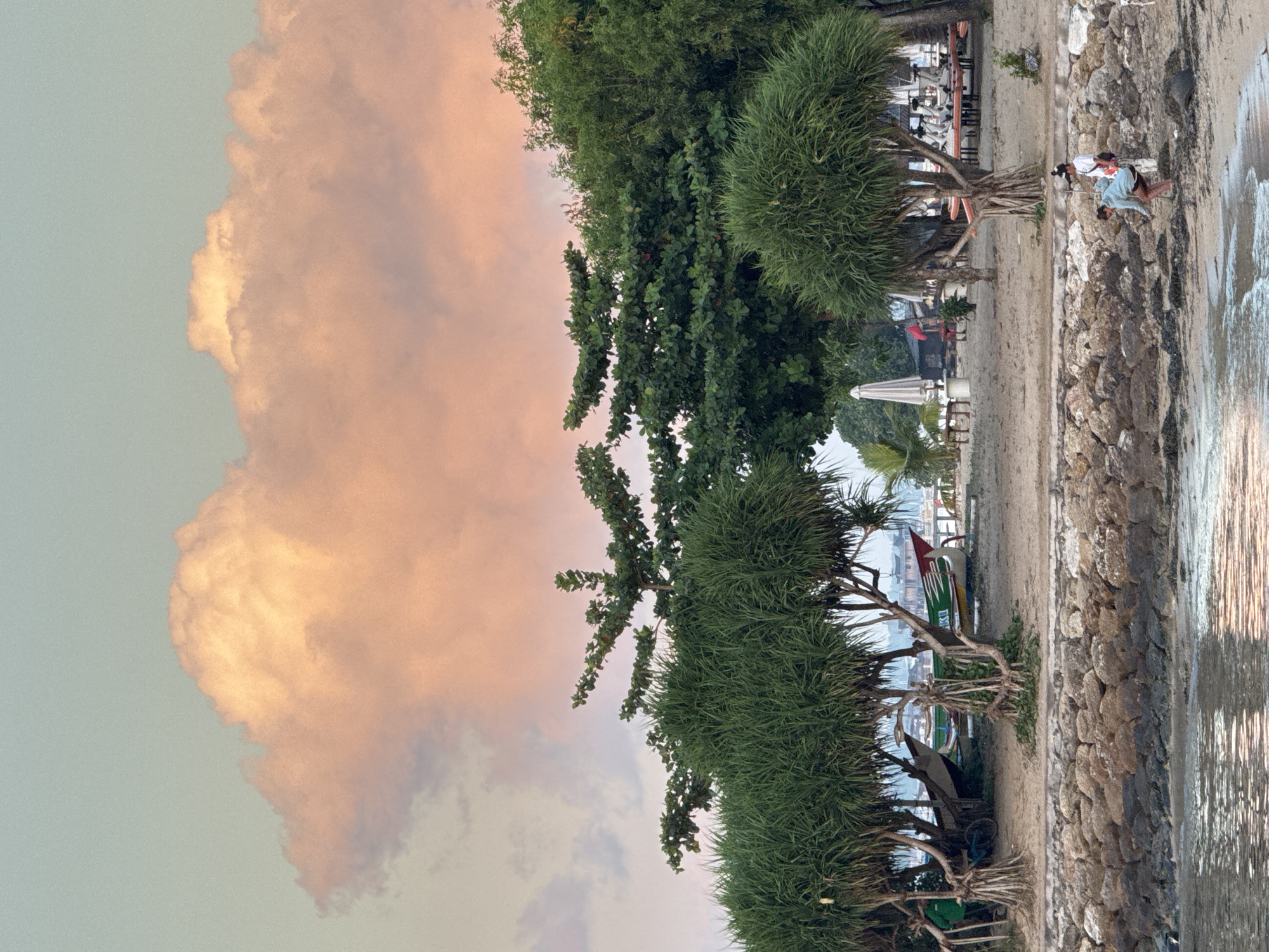 A wide-angle, eye-level shot captures a tranquil beach scene in Denpasar, Indonesia, with the sky dominated by a large, soft pink cloud. The foreground features the calm, rippling surface of the ocean, reflecting the muted light of dawn or dusk. A rocky embankment separates the water from the sandy shore, where two people are visible. One person, wearing a light blue hooded jacket and dark shorts, is crouched down, appearing to examine something on the beach. Another person, in a white top and dark shorts, stands nearby with a white bag slung over their shoulder.
Behind them, a line of lush green pandanus trees and other tropical foliage forms a natural barrier. Further back, several traditional Indonesian fishing boats with colorful decorations are beached on the sand. In the distance, glimpses of buildings suggest the proximity of a settlement. The overall atmosphere is serene and picturesque, with the soft colors of the sky and the gentle natural elements creating a peaceful ambiance.