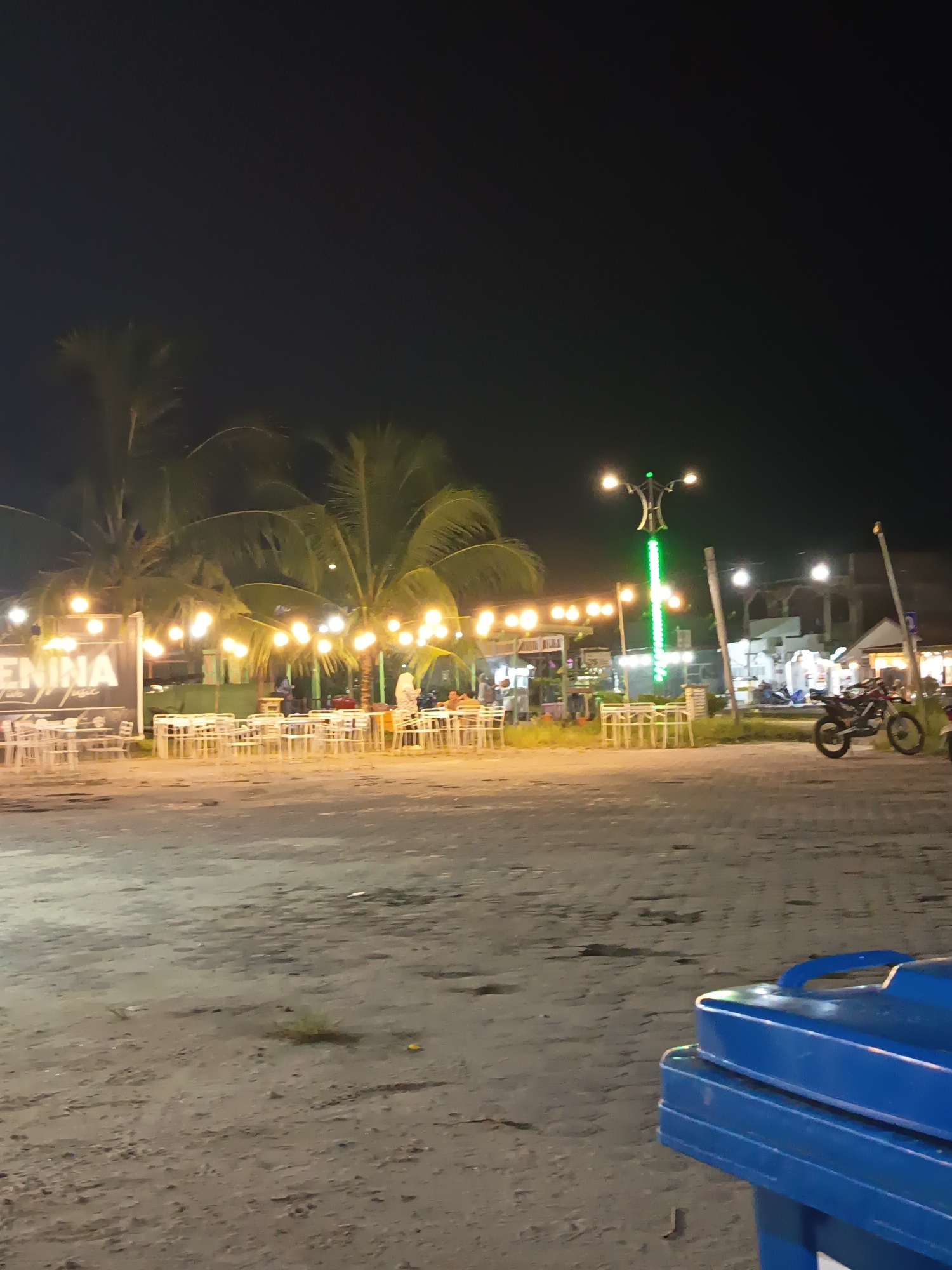 This image captures a nighttime scene at a beachfront establishment in Singkil, Indonesia, possibly a restaurant or cafe, illuminated by numerous string lights and a decorative street lamp. Palm trees are visible in the background. Several tables and chairs are set up on the sandy ground, suggesting an outdoor dining area. A motorcycle is parked to the right of the frame. In the foreground, a blue trash bin is positioned. The overall ambiance is one of a lively, illuminated outdoor venue at night.