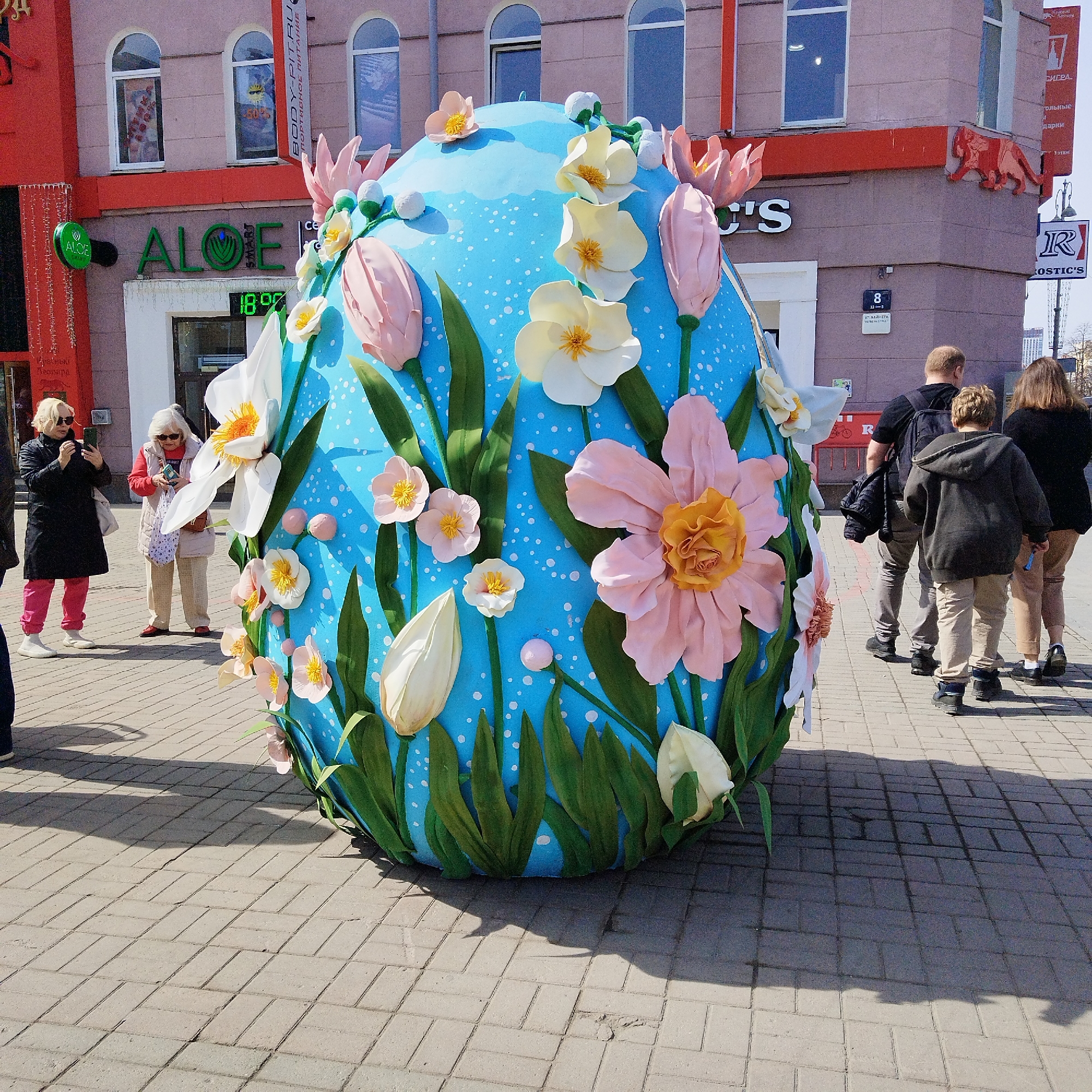 A large, blue, egg-shaped sculpture, intricately decorated with numerous pink, white, and yellow artificial flowers and green leaves, stands prominently in the foreground of a sunny urban street scene. The egg is covered in small white dots, adding to its decorative appeal. Its base is hidden by more green foliage.
Several people are observing the oversized egg, suggesting it is a public art display or seasonal decoration, likely for Easter. On the left, two older women are engaged with the egg; one is capturing a photo with her smartphone, while the other looks down, possibly at a mobile device or something in her hand. On the right side, a small group consisting of a man, a boy, and a woman walks away from the egg, their backs turned to the viewer. Strong shadows cast on the light-colored paving stones indicate bright daylight.
In the background, a light pink/purple building with arched windows features prominent red elements, including window frames and parts of the facade. Several signs are visible:
* "ALOE SMART" in green lettering with a matching green circular logo, below which a digital clock displays "1890".
* A vertical banner reads "BODY-PIT.RU" and "СПОРТИВНОЕ ПИТАНИЕ" (Sports Nutrition).
* A red sign with "ROSTIC'S" is visible on the right.
* Other Russian text includes "Красный Перицара" (Red Tsar), "УЛ. ВАЙНЕРА" (Vaynera Street) next to building number "8", and fragments like "СИГРА", "кольные", "дарки", and "Зэтаж". An embossed red lion or mythical creature is also visible on the corner of the building.
The scene depicts a vibrant street environment where passersby interact with a festive, large-scale art installation.