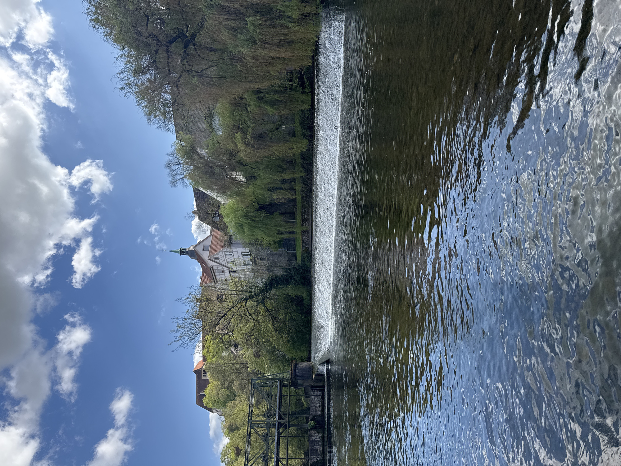 The image depicts the idyllic landscape around the Murr River in Backnang, Germany. The focus is on a small weir or dam that creates a gentle waterfall effect. Water flows over the weir into the river's expanse, reflecting the sky. On the left bank, greenery flourishes with trees lining the riverbank. A building with a distinctive spire rises above the trees. An old watermill mechanism, partially submerged, stands to the left of the weir. The sky is blue with scattered white clouds, suggesting a clear day. There are no people visible in the image.