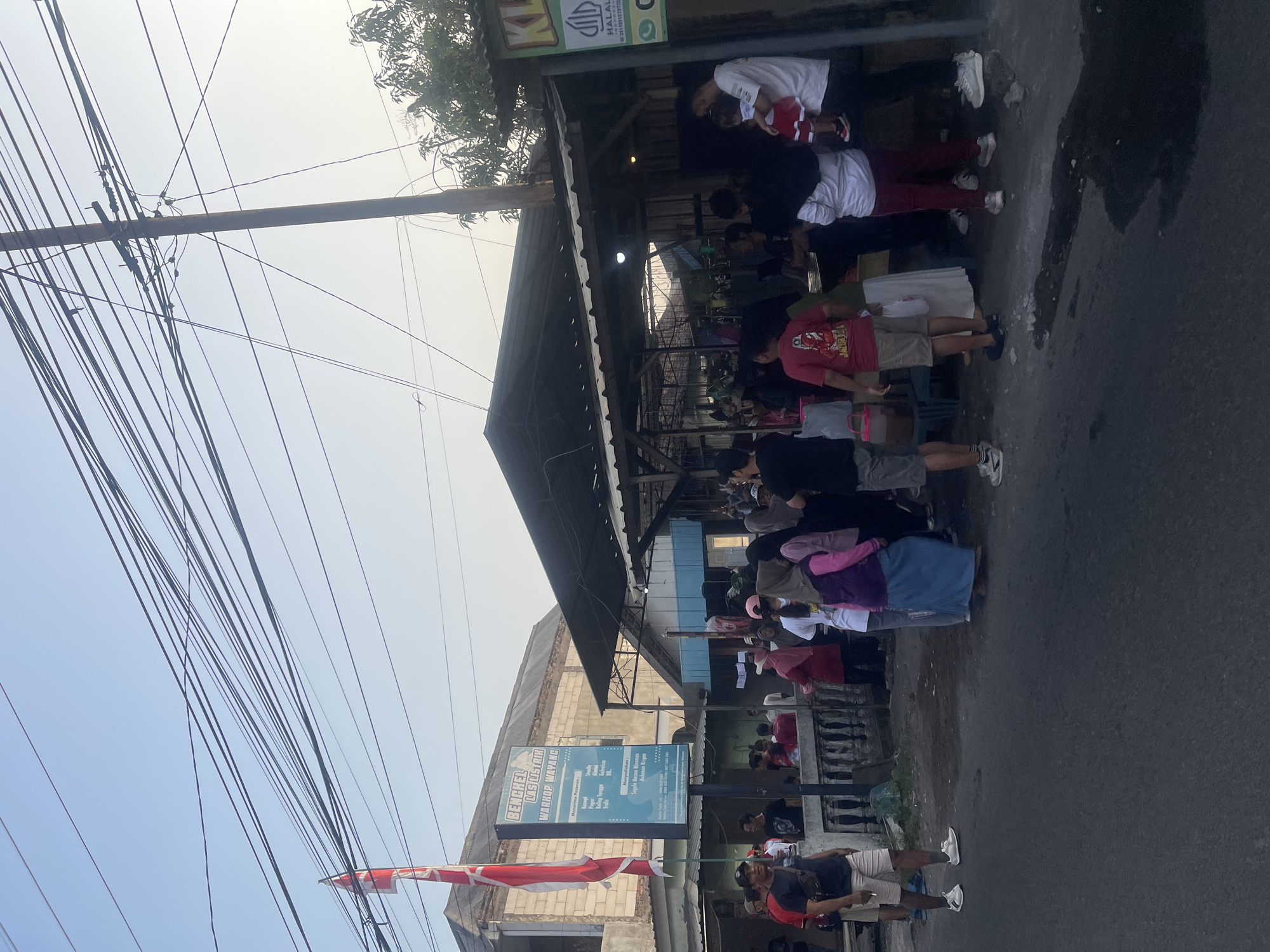 The image depicts a street scene in Kedungkendo, Indonesia, possibly during the late afternoon or early evening, given the lighting. A group of people are gathered around an outdoor food stall or small eatery. There is a building in the background with a sign that reads "BENGKEL LAS LISTRIK WARROP WAYANG". The stall has a corrugated metal roof supported by poles. Various individuals, including men, women, and a child being held by an adult, are seen standing, talking, and queuing. Some people are dressed in casual clothing, while others are wearing traditional head coverings. The ground in front of the stall is paved, with some puddles of water visible. Overhead power lines crisscross the sky, typical of an urban or semi-urban setting. A red and white flag is visible on a pole to the left of the frame. The overall impression is of a bustling local gathering place.