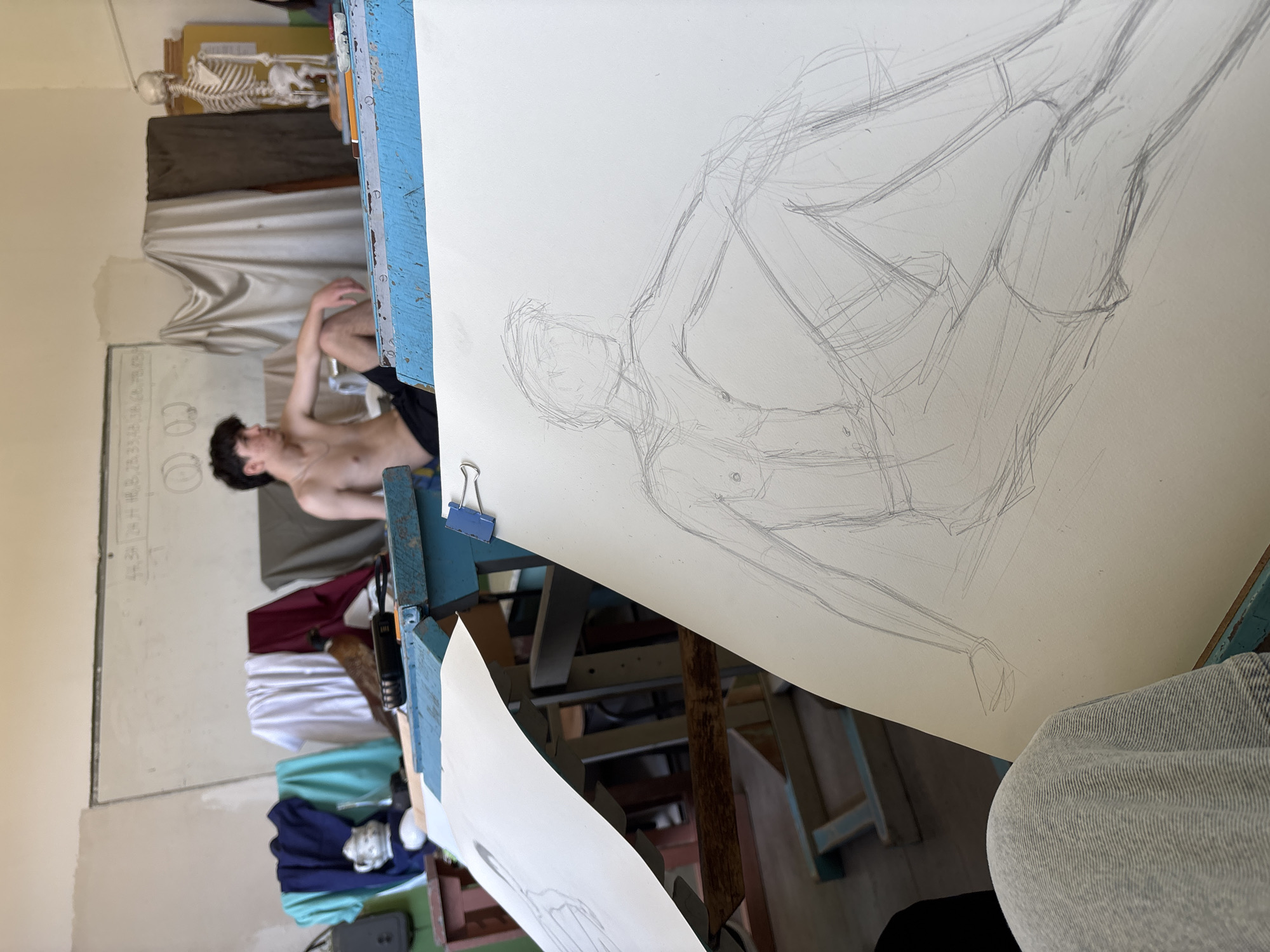 This image depicts a figure drawing session taking place in an art studio in Troyan, Bulgaria. The foreground is dominated by a large sheet of white paper on a blue, well-worn wooden easel, featuring a loose pencil sketch of a reclining male figure. The sketch, rendered with gestural lines, captures the pose of the live model visible in the background.
The model, a young man with dark, curly hair, is shirtless and reclines on a surface, his left arm bent and resting near his head, and his legs also bent. His relaxed posture is the subject of the artwork.
The studio environment behind the model includes a whiteboard with faint, illegible diagrams and text. Below the whiteboard and the model, an assortment of folded clothes in various colors (burgundy, white, turquoise, dark blue) is stacked. In the upper background, a full human skeleton model stands on a shelf, serving as an anatomical reference. Next to the skeleton, a draped piece of light fabric is arranged on a stand.
A blue binder clip secures the drawing to the board. The lower right corner shows a partial view of the artist's lap, clad in light-colored fabric, indicating their active participation. The scene conveys a focused, academic atmosphere typical of an art classroom, likely during daytime given the bright indoor lighting.