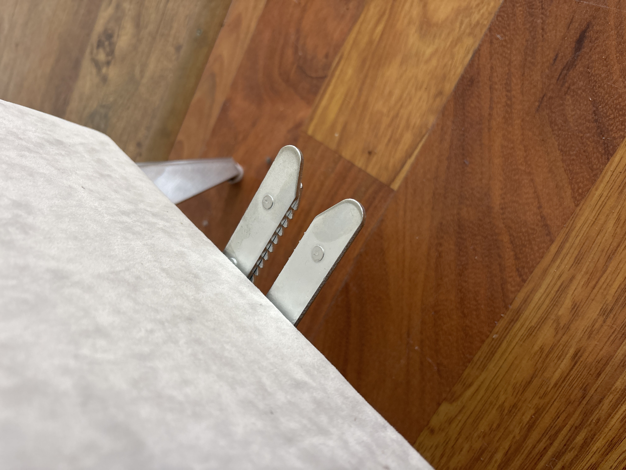 This is a close-up, eye-level shot of two silver-colored metal piano pedals. The pedals are positioned on a polished wooden floor with a warm, reddish-brown hue. The pedals are partially obscured by a piece of light-colored fabric, possibly upholstery from a piano bench or a section of the piano itself, which is at the top left of the frame. The fabric appears soft and textured. The pedals themselves have a serrated edge, suggesting they are designed for grip. One pedal is slightly above and in front of the other. The overall lighting is soft and diffused, casting subtle shadows. No people are visible in the image, nor is there any specific indication of the city of Zaporizhzhia, Ukraine. The scene appears to be an interior shot focused on the details of the piano pedals and flooring.