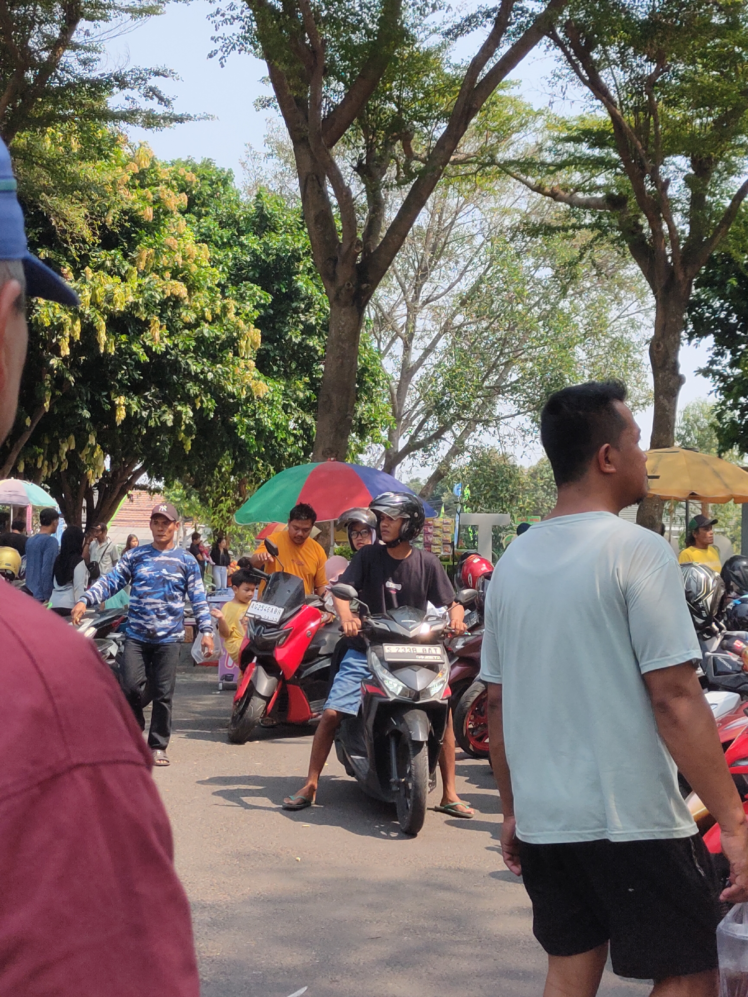 A medium shot captures a street scene in Ngasem, Indonesia, under a bright, clear sky. Several people are present, mostly engaged in activities related to motorcycles. In the foreground, a person wearing a maroon shirt is partially visible on the left, facing away from the camera. To their right, a man in a blue and black camouflage patterned long-sleeved shirt and black pants walks forward, appearing to gesture or lead. He wears a black baseball cap.
Further into the scene, a young child in a yellow shirt stands near a red and black motorcycle. Behind the child, a man in an orange shirt is positioned on the left side of a motorcycle, looking towards the child. Another individual, wearing a black t-shirt and a helmet, sits on a motorcycle in the center, facing slightly to the right. To the right of the frame, a man in a light blue t-shirt and black shorts is seen from the back, looking to the right. He is holding a clear plastic bag in his right hand.
Several motorcycles are parked or in motion, with one red and black motorcycle and a dark colored motorcycle prominently featured. Colorful umbrellas are visible, one red, green, and blue, and another pink and white, providing shade. Trees with green foliage and some with yellow blossoms line the street, and a yellow awning is visible in the background. The ground is asphalt. The overall atmosphere appears to be that of a public gathering or marketplace.