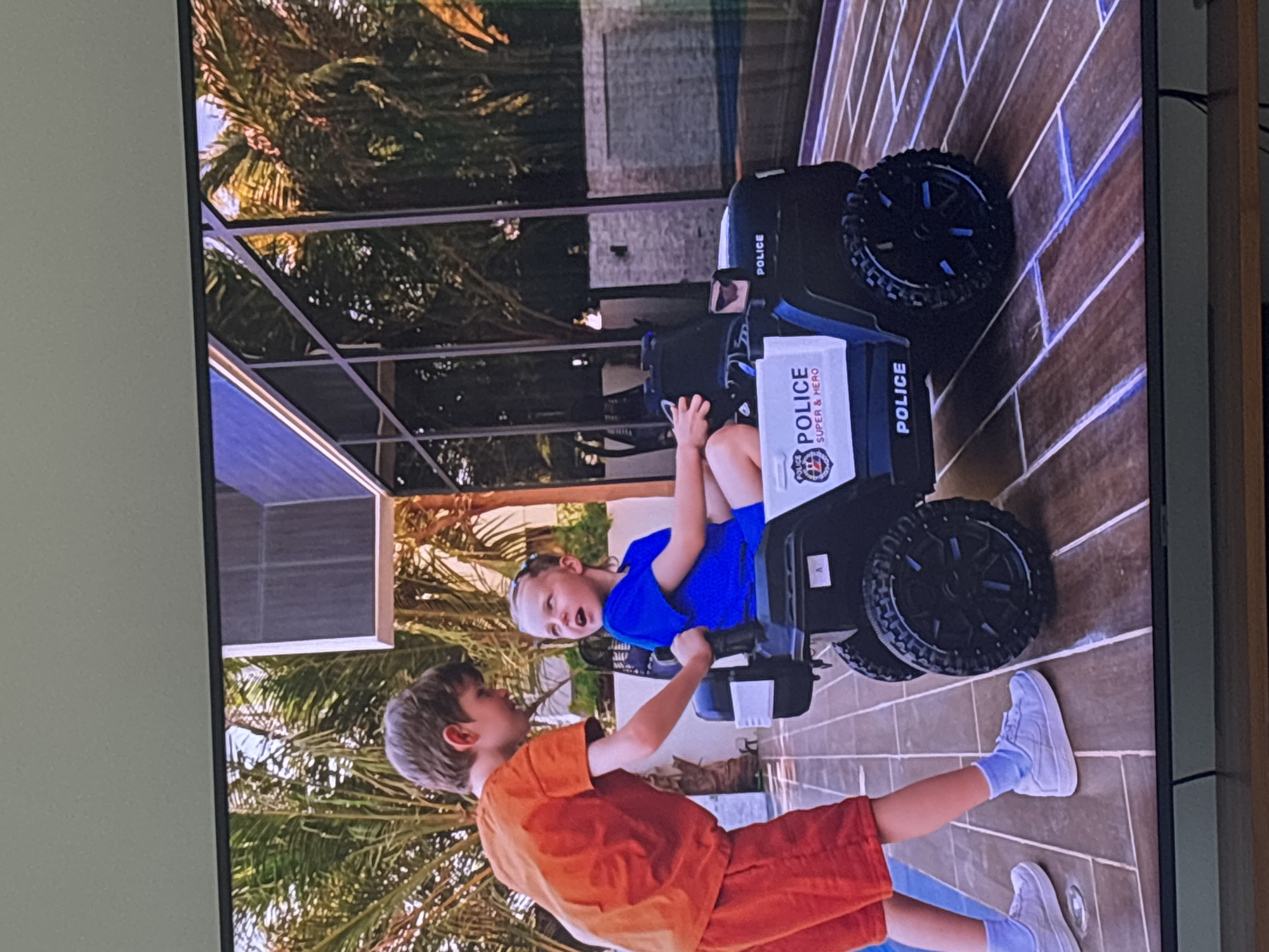 Two young boys are interacting outdoors on a tiled patio. One boy, wearing an orange t-shirt and orange shorts, is standing and looking down at the other boy. The second boy, wearing a blue t-shirt and blue shorts, is sitting in a black and white toy police car. The police car has "POLICE SUPER & HERO" printed on its side. Palm trees and modern architecture, including large windows and what appears to be a swimming pool, are visible in the background, suggesting a warm climate. The setting is consistent with a luxury residential area in Dubai, United Arab Emirates, where such outdoor spaces and amenities are common. The lighting suggests it is daytime.