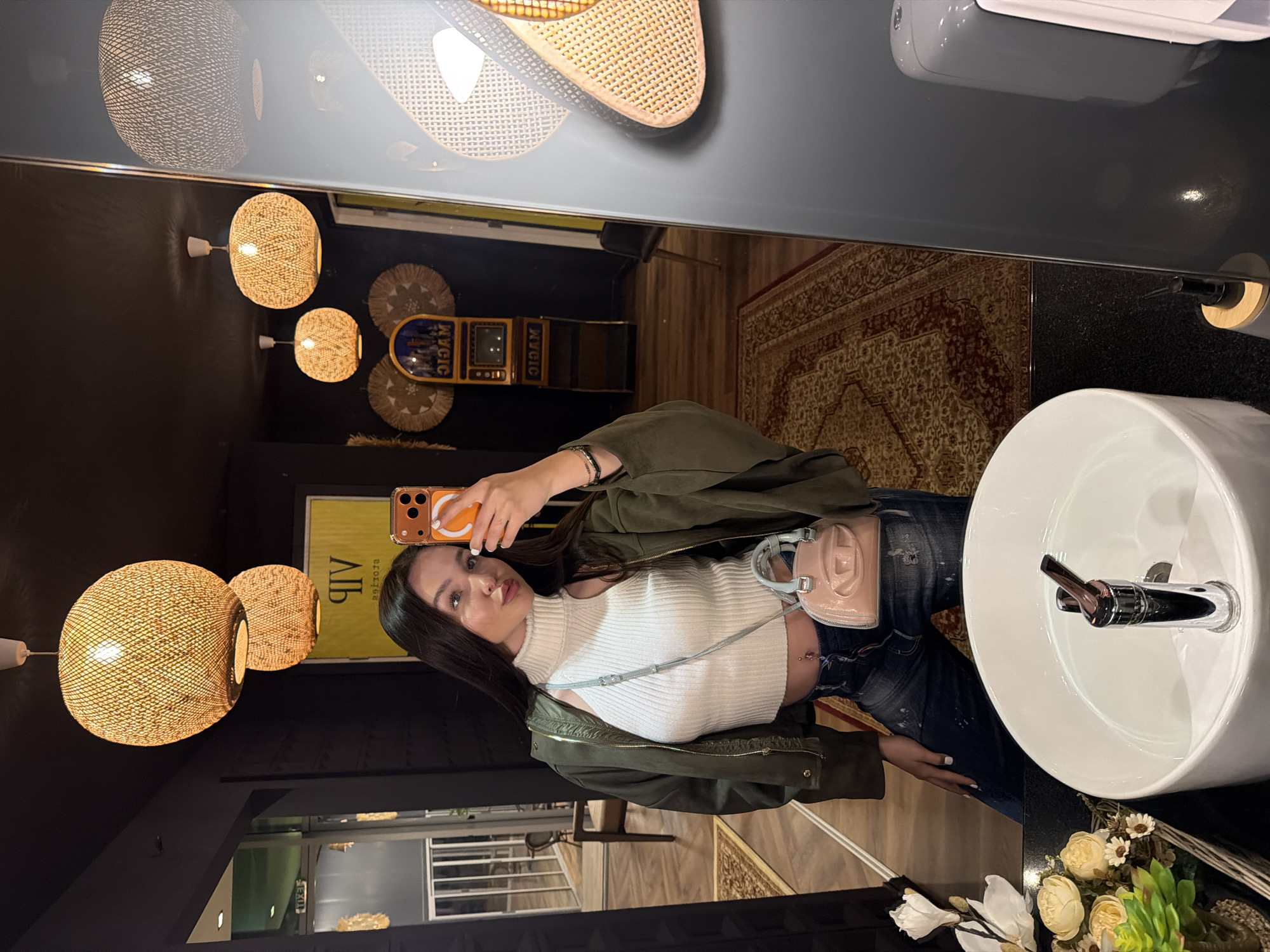 A woman is taking a selfie in a bathroom. The bathroom has a round sink, black countertop, and decorative flowers. Wicker light fixtures are visible in the mirror. The background shows a rug and a game machine. There is no indication of the location being Kulata, Bulgaria.