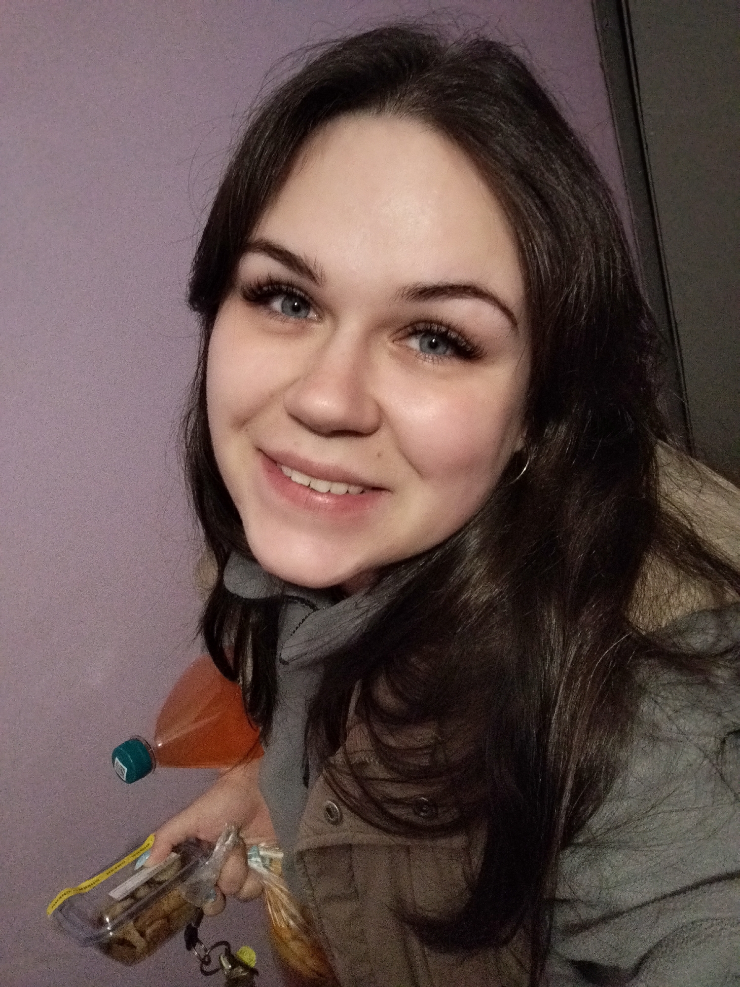 The image shows a young adult female, present in an indoor setting within the city of Chaikovsky, Russia. She is positioned centrally, facing the camera with a slight smile. She has long, dark brown hair and blue eyes with prominent eyelashes. A small silver hoop earring is visible in her left ear. She is wearing a grey hooded garment beneath a light brown or tan jacket, which features a beige fur-like trim visible near her right shoulder. In her right hand, located in the lower left portion of the frame, she holds a transparent plastic container containing light-colored items, possibly food, and a set of keys on a keyring. An orange plastic bottle with a green cap, containing an orange liquid, is visible behind her hand. The background consists of a plain light purple wall, with a dark vertical structure, likely a door frame or cabinet edge, positioned on the far right.