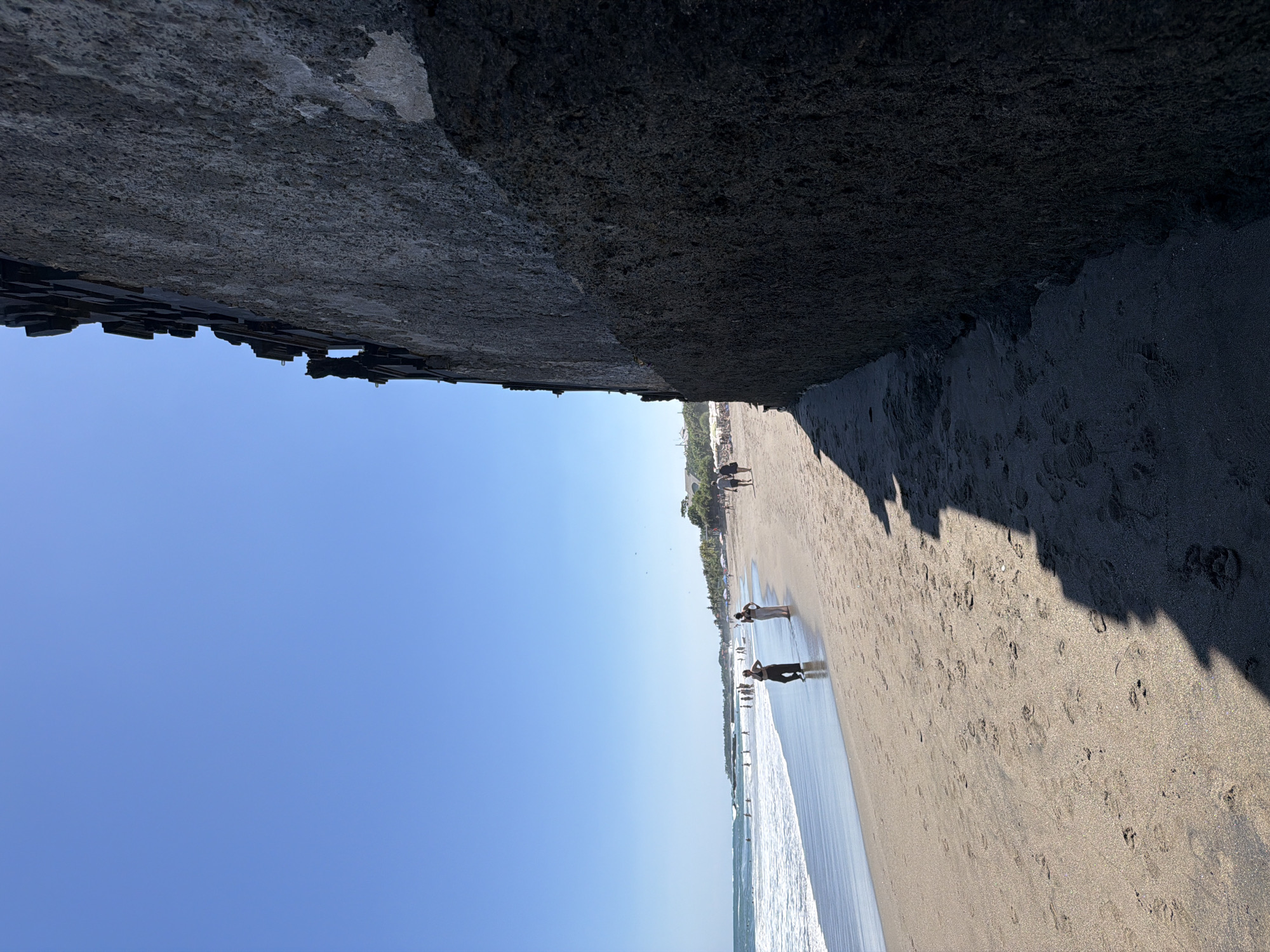 The image depicts a beach scene in North Kuta, Indonesia, from a low-angle perspective. The top of the image features a dark, coarse concrete structure, possibly a seawall or the base of a building. Below this, the scene transitions to a sandy beach with a visible waterline where gentle waves are breaking. The sand is light-colored and appears damp near the water. Several people are walking along the shoreline, their forms somewhat obscured by the angle and distance. Beyond the immediate foreground, a row of trees and buildings lines the edge of the beach. The sky is clear and bright blue, suggesting a sunny day. Shadows cast by the concrete structure and possibly by the people on the beach are present on the sand.