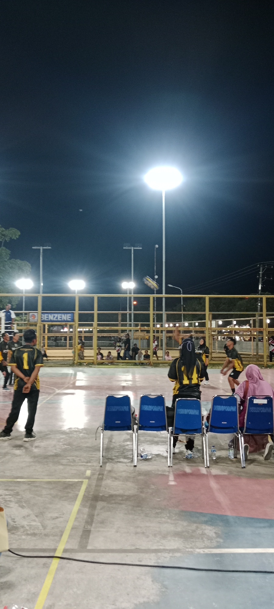 A volleyball game is underway at night under bright floodlights in Tuban, Indonesia. The court is concrete with colored markings. Spectators are seated on blue chairs, some wearing headscarves. Players in matching yellow and black uniforms are actively participating in the game. A banner with the word "BENZENE" is visible behind the fence, and the surrounding area appears to be an outdoor sports facility.