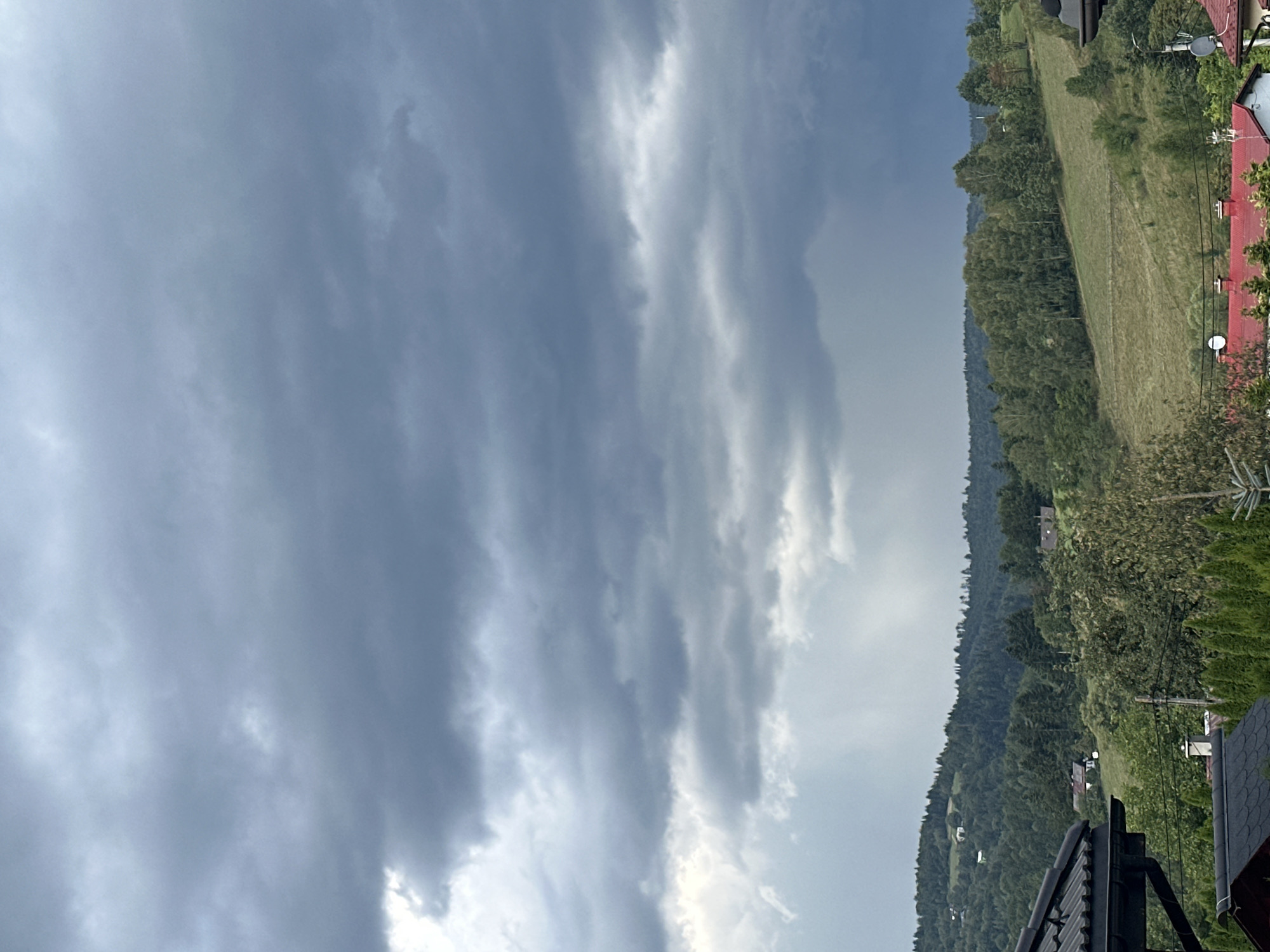 The image depicts a cloudy sky over a hilly, forested landscape with scattered houses and rooftops in the foreground. The sky is filled with heavy, dark grey clouds, suggesting an impending storm or rain. Below the sky, a green hillside is visible, dotted with trees of various shades of green. Several houses with red and grey roofs are nestled among the trees and on the slopes. In the immediate foreground, parts of two house roofs are visible, one with dark grey tiles and the other with red tiles. A few coniferous trees are also present in the foreground. The scene appears to be a rural or semi-rural area, consistent with the outskirts of Wisła, Poland, known for its hilly terrain and forested natural beauty. No people are visible in the image.