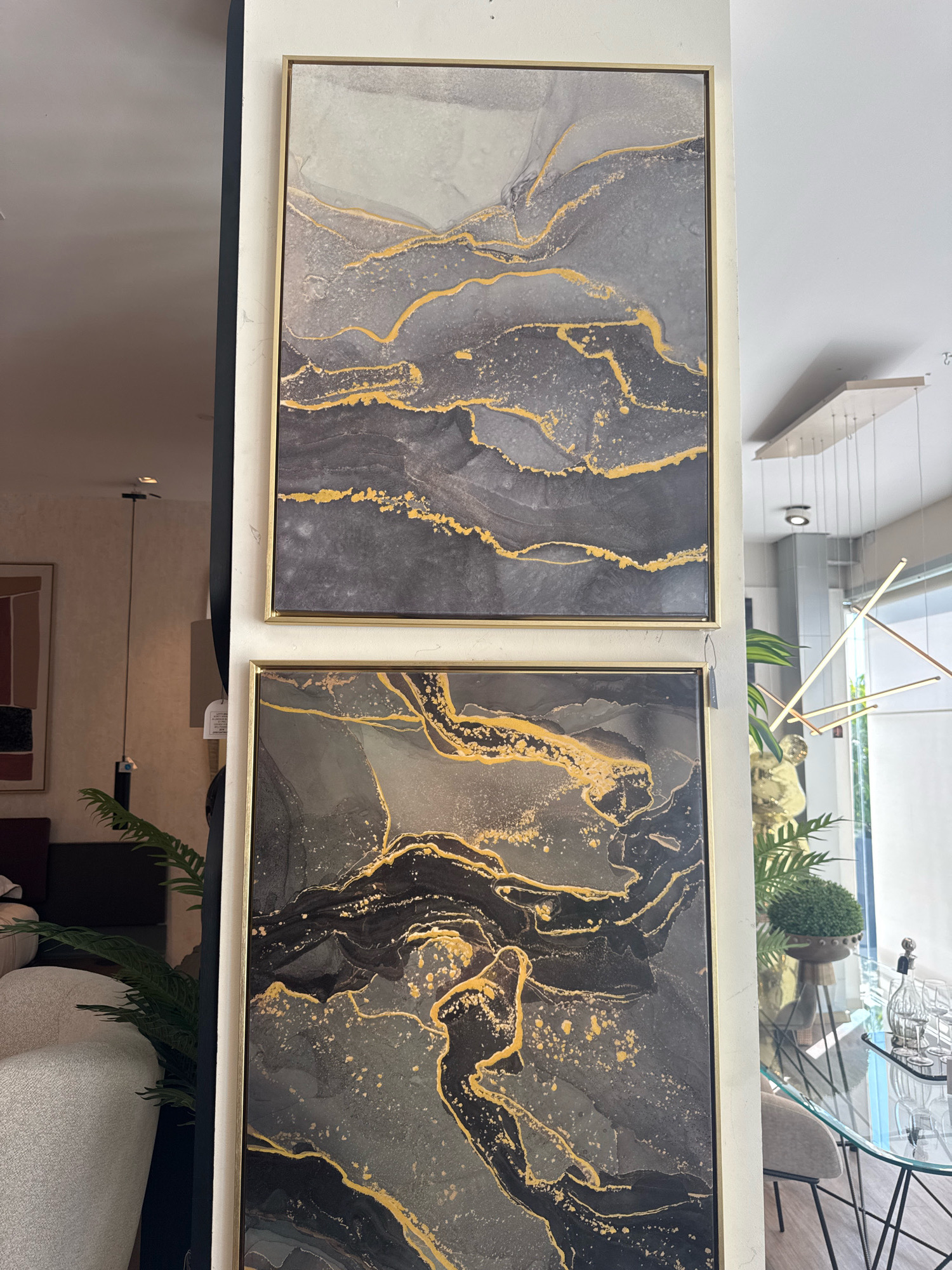 This is a close-up, eye-level, indoor shot of two framed abstract paintings with gold veins, displayed on a white wall in a store. The paintings are arranged vertically, with the top painting featuring swirls of gray and white with scattered gold accents. The bottom painting is similar in style but with darker, more prominent black and gray areas and more extensive gold veins. The frames appear to be a thin gold color. To the left of the paintings, a dark pillar or wall casts a shadow. In the background, a lush green plant is visible, and further back, a glass-top table with chairs can be seen, along with a large mirror reflecting the environment. In the upper right, a modern light fixture with thin metallic rods hangs from the ceiling. This scene is likely from a furniture or home decor store located in the Agios Athanasios Municipality, Cyprus, suggested by the tag on the bottom painting, which is in Greek.