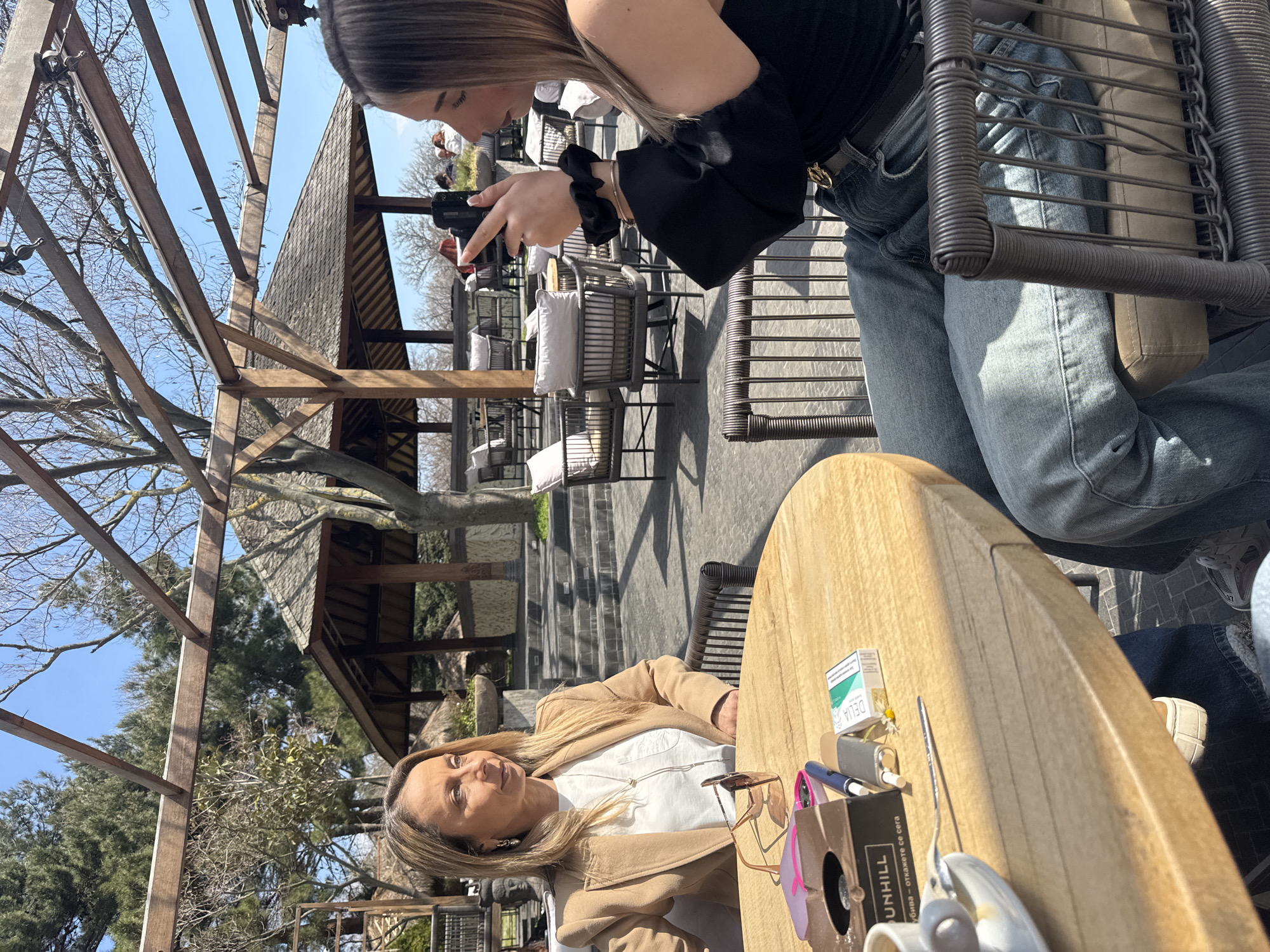 A woman sits at an outdoor table at the SUNHILL restaurant in Varna, Bulgaria. She wears a tan blazer and a white shirt. Another woman leans over the table, holding a camera, presumably to take a photo of her. A pack of DELIA cigarettes, sunglasses, a lighter, a pen, and other personal items are on the table. The restaurant features an outdoor seating area with a wooden pergola and views of lush greenery. Other patrons and seating arrangements can be seen in the background.