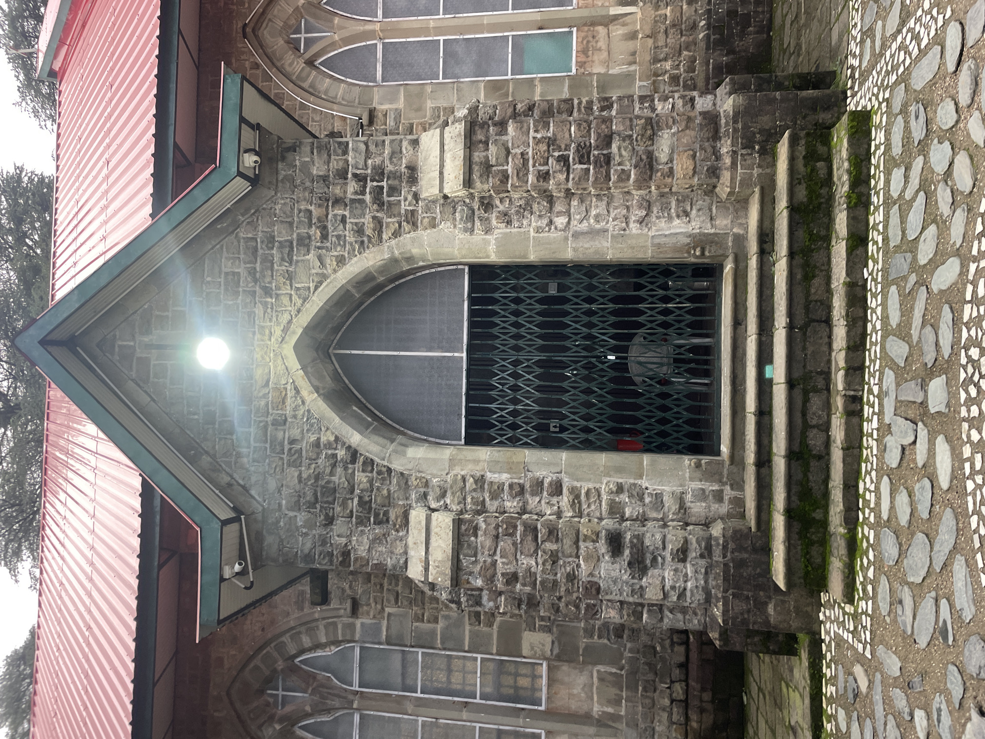 The image shows the entrance of a stone building, likely a church, in Dharamkot, India. The building features a red, corrugated metal roof with a green trim. The facade is constructed of rough-hewn stone blocks. An arched doorway is covered by a green, collapsible metal gate. Above the doorway, a prominent spotlight illuminates the entrance. Two security cameras are visible under the eaves of the roof. Stone steps lead up to the entrance, and the ground in front is paved with irregular stones and white decorative tiles. The windows on either side of the entrance are tall and arched, with leaded glass panes. The overall scene suggests an older, established building in a potentially damp environment, as evidenced by moss growing on the steps. The background shows trees, indicating a natural setting.