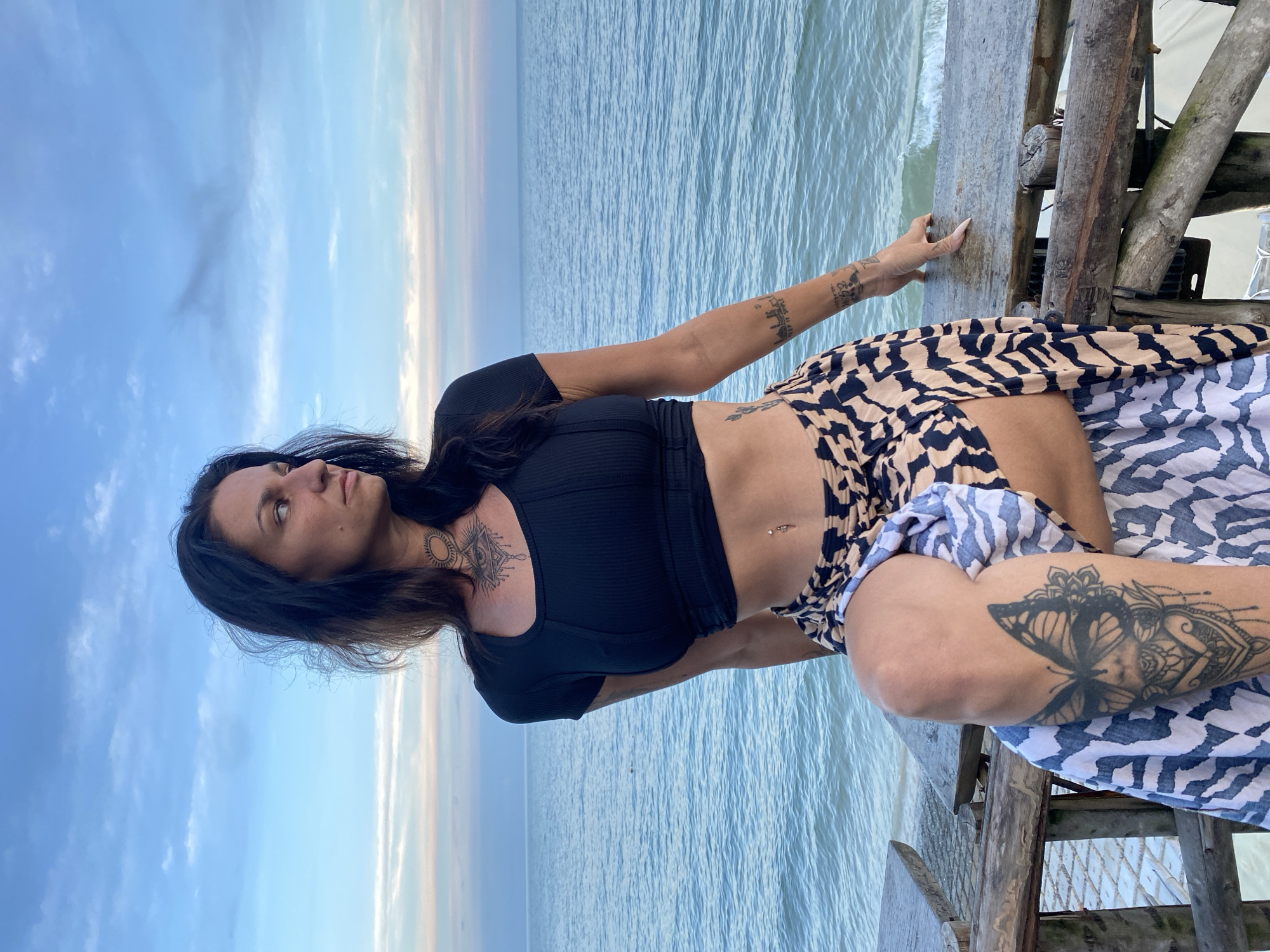 A medium shot captures a woman with dark hair and tattoos posed on a wooden railing with the ocean and sky in the background. The woman is wearing a black crop top and a patterned skirt. Her left leg is bent, with a butterfly tattoo prominently displayed on her thigh. The ocean is calm, with gentle waves. The sky is a gradient of blue and pink, suggesting either sunrise or sunset. The wooden railing appears weathered, indicating an outdoor setting, possibly a pier or boardwalk. The location is described as Phu Thuy Ward, Vietnam.