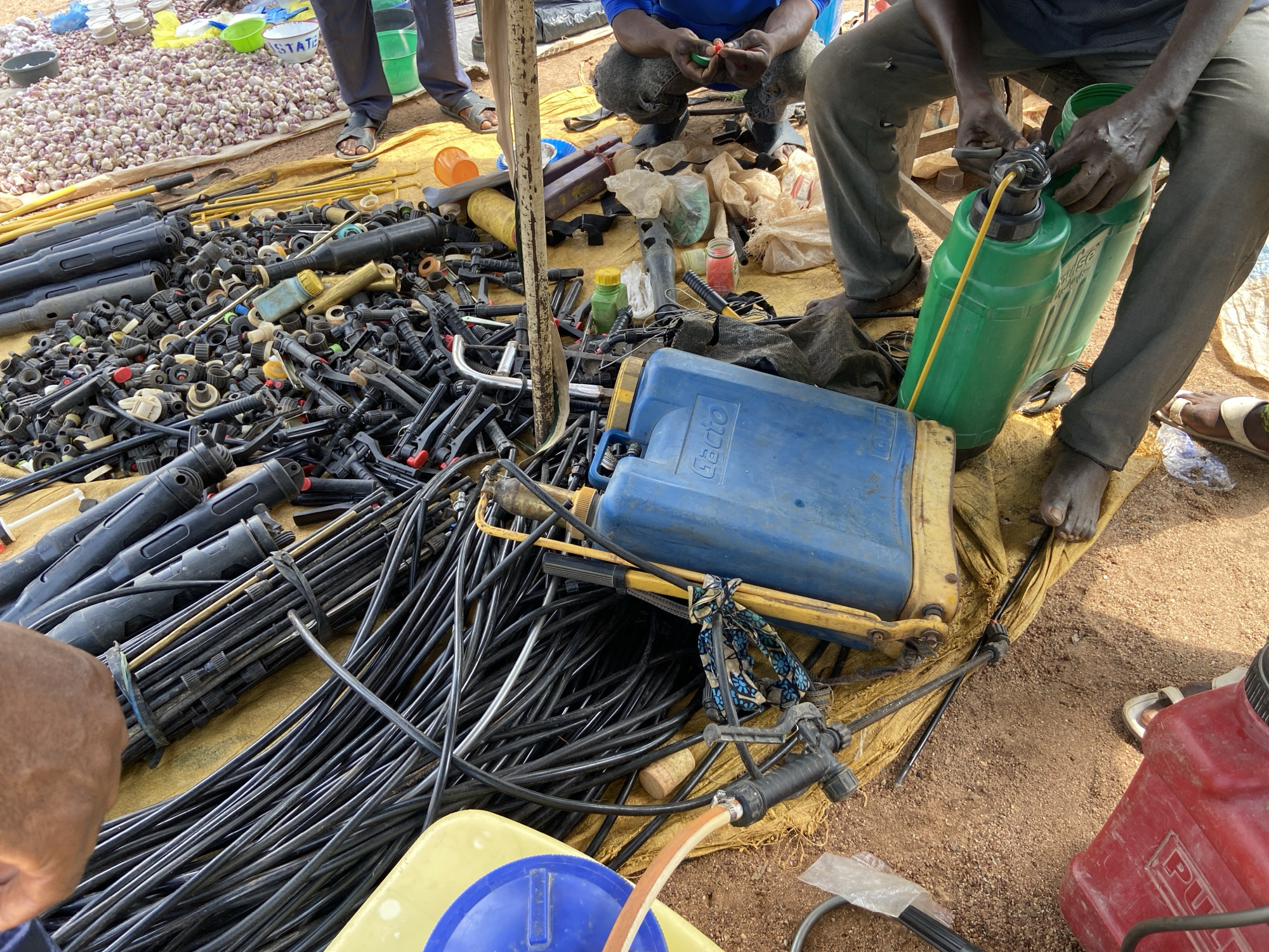 This image captures a busy outdoor scene, likely at an agricultural market or supply stall in Mararaba, Nigeria. The foreground and center are dominated by a large collection of agricultural equipment spread across a yellow tarp laid on sandy ground.
Several men are present and engaged in various tasks. A man in grey pants, barefoot, is prominently featured on the right, actively working with a green manual chemical sprayer. His hands are busy with its mechanism, suggesting he might be assembling, repairing, or filling it. Another man, wearing a blue shirt, is crouched in the upper middle, also focused on small components. A third person's legs and feet, clad in sandals, are visible in the top left, standing beside a large pile of white garlic bulbs. A fourth individual's hand is visible in the bottom left, near bundles of hoses.
The yellow tarp holds an extensive assortment of black plastic agricultural components, including numerous spray nozzles, connectors, and parts for irrigation or spraying systems. Bundles of black hoses and tubes are stacked, along with several long black plastic spray wands. Among these tools are a blue manual sprayer with "Gactio" visible on its side, the green sprayer being handled, and a partially visible red sprayer in the bottom right, which shows "PUM..." on its label. Yellow plastic spray lances, various small plastic bottles (some green, some yellow, some clear, possibly containing chemicals), and plastic bags containing other items are also scattered across the tarp. Empty plastic buckets and an orange plastic cup are visible in the background. The bright natural light indicates it is daytime. A white bucket in the background shows part of the word "STATE". The scene conveys a practical and industrious environment focused on farming supplies.