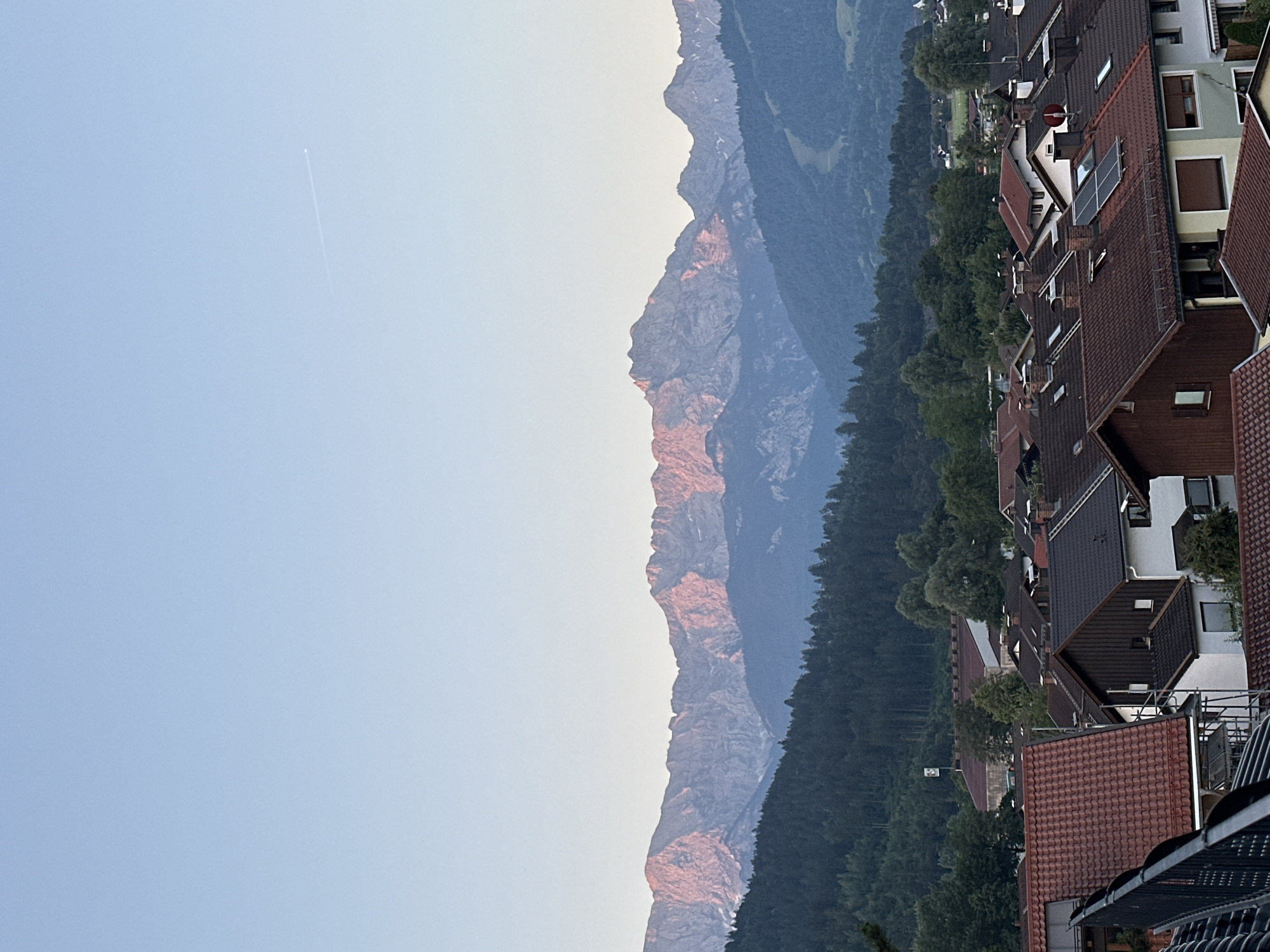 This is a photograph taken in Garmisch-Partenkirchen, Germany, featuring a panoramic view of the Alps at sunset. The sky is a clear, soft blue, with a faint airplane contrail visible high in the distance. The majestic mountain range dominates the upper half of the image, with its peaks illuminated by the warm, rosy glow of the setting sun. The mountains are rugged and imposing, with visible rock formations and snow-capped sections.
Below the mountains, a dense, dark green forest stretches across the landscape, creating a striking contrast with the illuminated peaks. Nestled within the foothills and extending into the foreground is a residential area characterized by numerous rooftops. These houses exhibit a variety of architectural styles, with dominant tile roofs in shades of brown and red. Some rooftops are equipped with solar panels, indicating a modern adaptation to the traditional housing. The overall impression is one of a serene, picturesque mountain town at the end of the day, with nature and human settlement coexisting harmoniously.