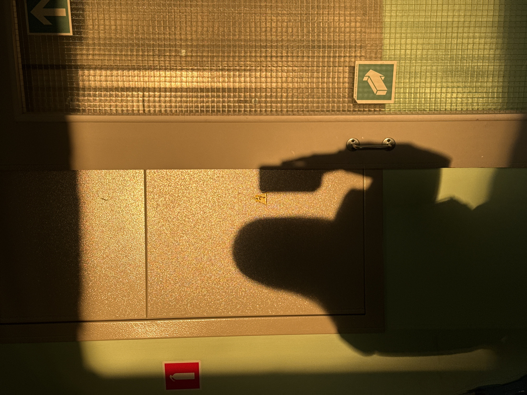 The scene depicts an indoor hallway or corridor in Chaikovsky, Russia, illuminated by strong, warm, golden light entering from the left. A large, dark shadow of a person, identified as the photographer, is prominently cast across a textured, golden-brown metallic utility cabinet or door and an adjacent pale green wall. The shadow distinctly shows the outline of a head, shoulders, and a hand holding a rectangular object, likely a smartphone, in the act of capturing the image.
On the green wall to the far left, a red square sign with a white fire extinguisher pictogram is affixed. The metallic cabinet/door structure, which exhibits a granular surface texture, features a small yellow triangular warning sign with a black exclamation mark on its upper panel. A rounded, metallic door handle is visible on the right edge of this structure. To the immediate right, a door or large panel made of textured, translucent glass, displaying a distinct grid pattern, reflects the intense golden light. Affixed to this patterned glass are two green square signs with white arrows: one positioned near the top right, pointing directly upwards, and another positioned near the bottom right, pointing diagonally right and upwards.