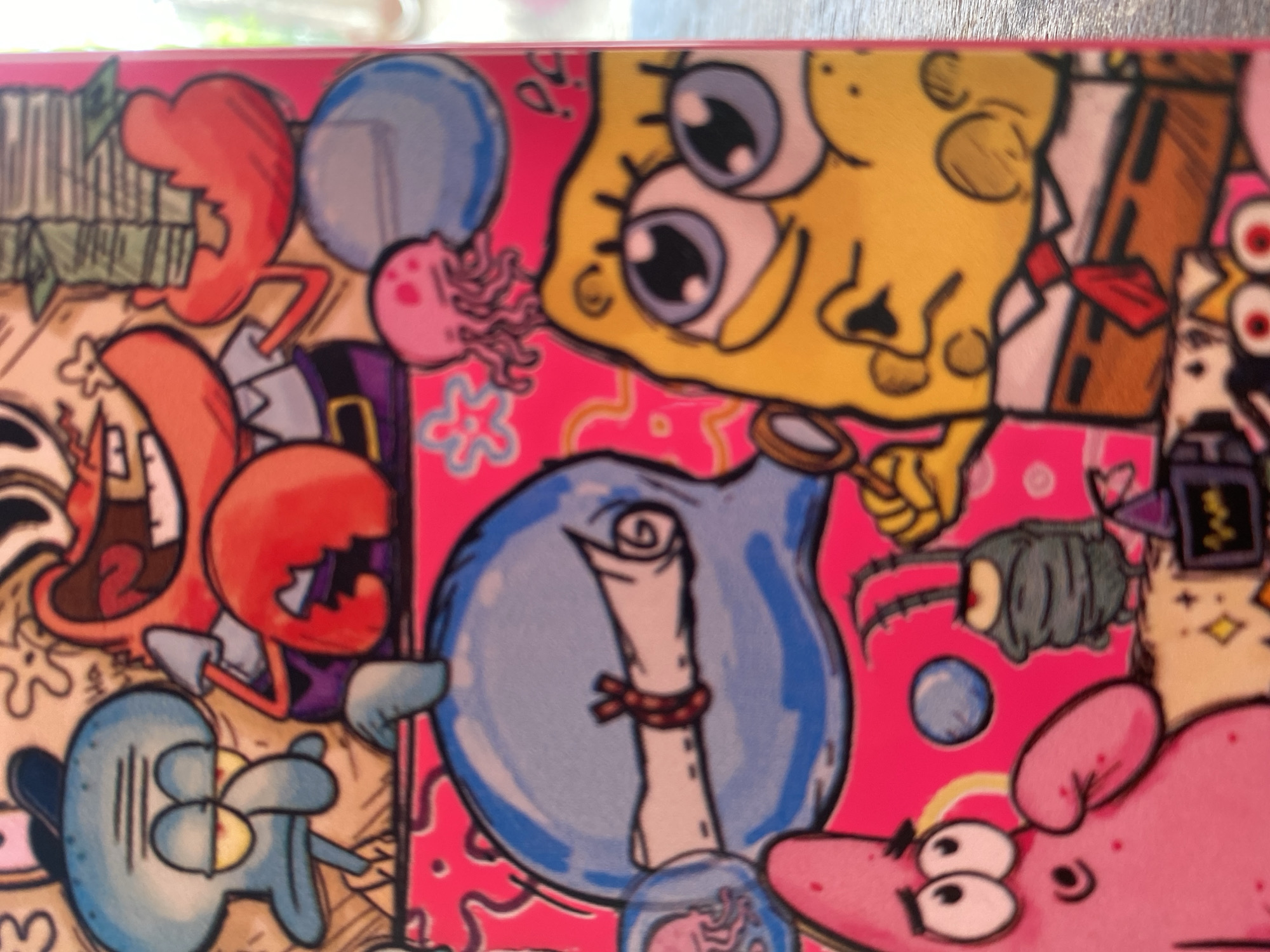 The image is a close-up, high-angle photograph of a vibrant, colorful illustration featuring characters from the cartoon SpongeBob SquarePants. The style is a dense, busy drawing with a pink background. In the foreground, SpongeBob is depicted with a magnified glass, looking upwards with wide eyes. To his left, Patrick Star is shown with a surprised expression, his arm extended. A blue jellyfish is floating above SpongeBob. In the middle ground, a large blue, ghost-like figure with a scroll tied with red ribbon is prominent. To the left of this figure, Squidward Tentacles is shown with a grumpy expression. Mr. Krabs, with his large claws, is to the right of Squidward, holding a stack of money. A pink jellyfish is also visible in this section. In the background, more background elements like stars and bubbles are visible. The image appears to be a still from a digital artwork or a printed item such as a phone case or poster. There is no indication of Ketileng, Indonesia, or any urban environment.