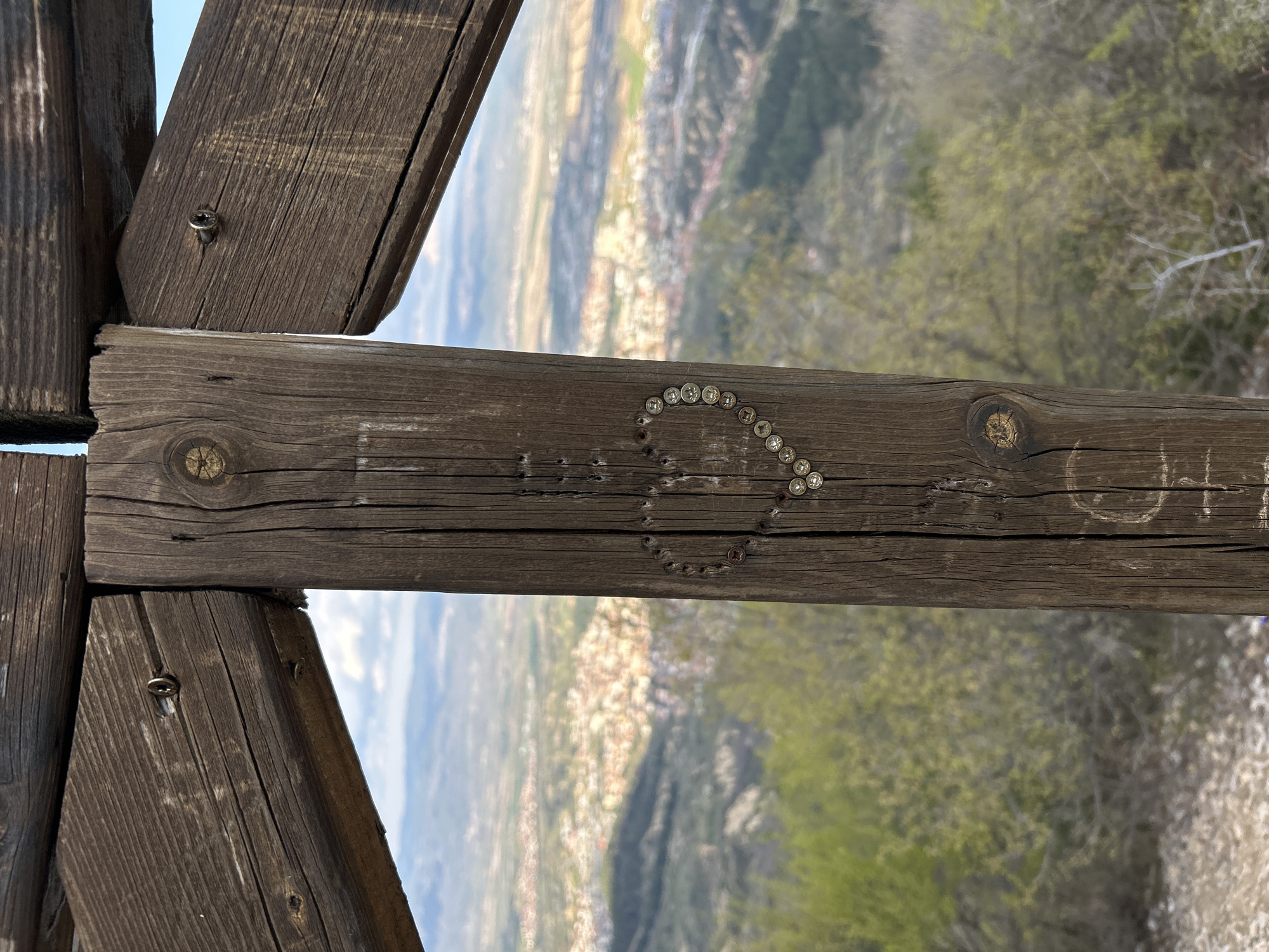 This image shows a weathered wooden crossbeam structure, likely part of an overlook or viewing platform. The crossbeam is decorated with a heart shape formed by numerous small, round, metallic objects, possibly screws or decorative studs. There are also carved inscriptions on the wood, with the letters "OH" faintly visible. The backdrop features a panoramic view of a city nestled in a valley surrounded by green hills and trees. Based on the prompt, the city is Delchevo, Bulgaria. The sky is partly visible in the upper left corner.