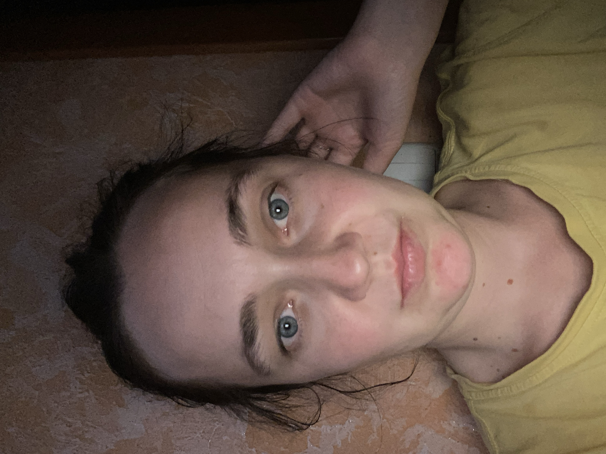 A frontal medium shot captures a young woman indoors, with a textured, light brown wall in the background. She has her hair pulled back, and her right hand is resting on the back of her head, partially obscuring her ear. The woman is wearing a yellow t-shirt. Her blue eyes are looking directly at the camera. The lighting is dim, with a warm cast, suggesting the presence of artificial light sources. The overall scene appears to be a personal, candid shot taken in a domestic setting.