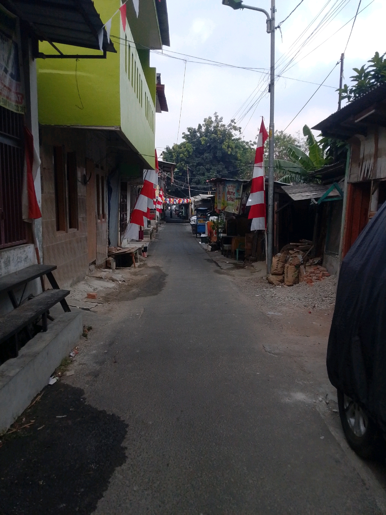 The image depicts a narrow street in East Jakarta, Indonesia, decorated with Indonesian flags, likely for a national holiday. The street is flanked by low-rise buildings, some of which are painted bright green. Red and white Indonesian flags are prominently displayed, hung from poles and buildings. Benches are visible along the left side of the street. The road surface is asphalt, and some debris is scattered along the sides. Several sacks of material, possibly cement or fertilizer, and stacks of bricks are piled up on the right side of the street, next to a building with a corrugated metal roof. Overhead, electrical wires crisscross the sky. A street lamp is visible on the right. In the background, trees and more buildings suggest a densely populated urban area. A vehicle covered with a dark tarp is parked on the right side of the road, with its wheel visible. The scene appears to be a typical residential or commercial street in a bustling Indonesian city.