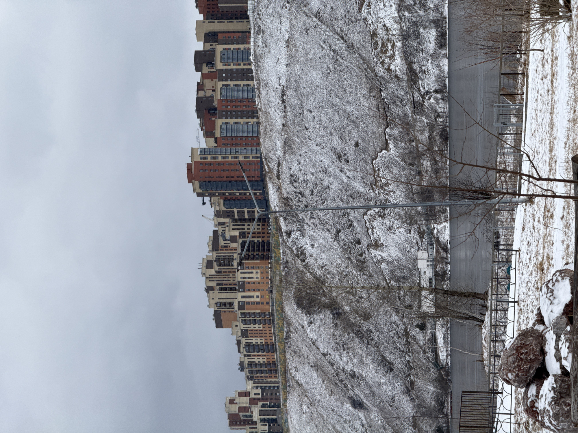 The image depicts a wintry scene in Krasnoyarsk, Russia, featuring a row of modern apartment buildings along the left edge. The buildings are of varied heights and colors, mainly in shades of brown, tan, and brick red. To the right of the buildings, a snow-covered hillside slopes down towards what appears to be a body of water, possibly the Yenisey River. The hillside is largely barren, with sparse vegetation visible under the snow. In the foreground, there is a metal fence and a cluster of snow-covered rocks. A few bare trees are scattered across the scene. The sky is overcast, suggesting a cold and gloomy winter day.