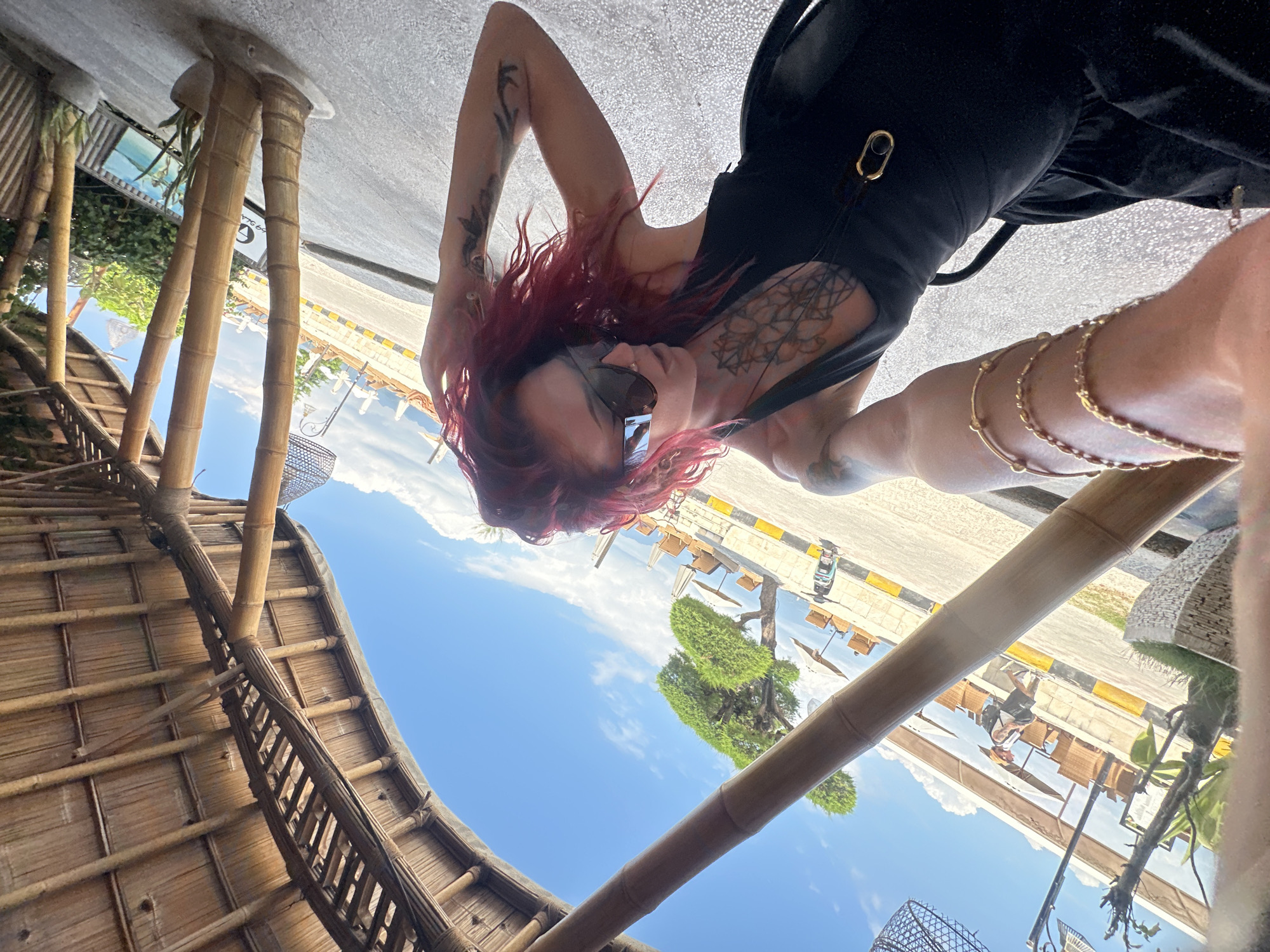 A rotated image shows a person with red hair and tattoos on their arms and chest wearing a black outfit and sunglasses. The background includes a bamboo structure, a blue sky with clouds, trees, and what appears to be a road with yellow and black markings. Further in the background, there are buildings and other structures. The location is Unknown, Indonesia.