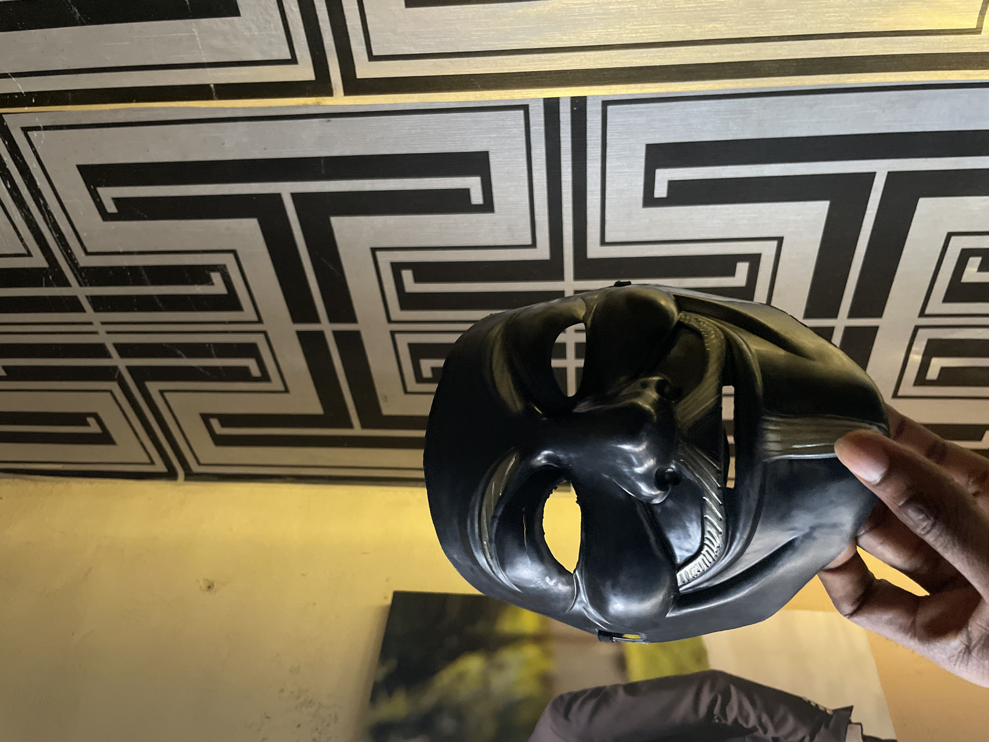 A black mask is held up by a dark-skinned hand in front of a wall with a geometric pattern of black and silver. Below the mask is a tan-colored surface. Below that, out of focus, is part of a picture or another object. The scene is indoors with fluorescent lighting. The location is not identified. The city of Lokoja, Nigeria, is not mentioned in the scene.