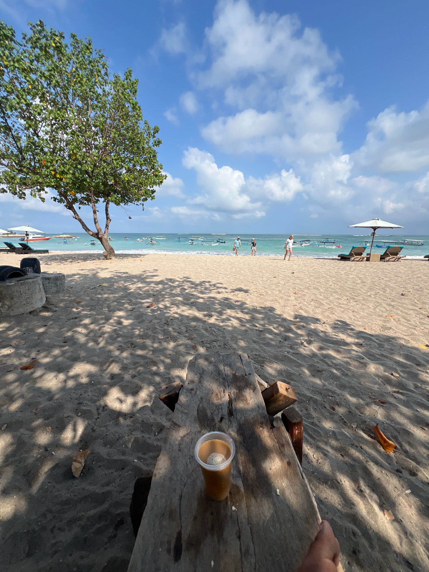 A perspective shot from a wooden bench on a sandy beach in Kuta, Indonesia, shows a plastic cup of coffee with foam on a rough-hewn wooden table in the foreground. Beyond the bench, the beach stretches towards the turquoise ocean, dotted with traditional Indonesian jukung boats. In the distance, several people are visible walking along the shoreline or wading in the shallow water. Two sun loungers are placed under a white umbrella, and a large green tree with a visible trunk casts shadows across the sand. The sky is a bright blue with scattered white clouds, indicating a sunny day. The overall scene depicts a relaxed beach environment.