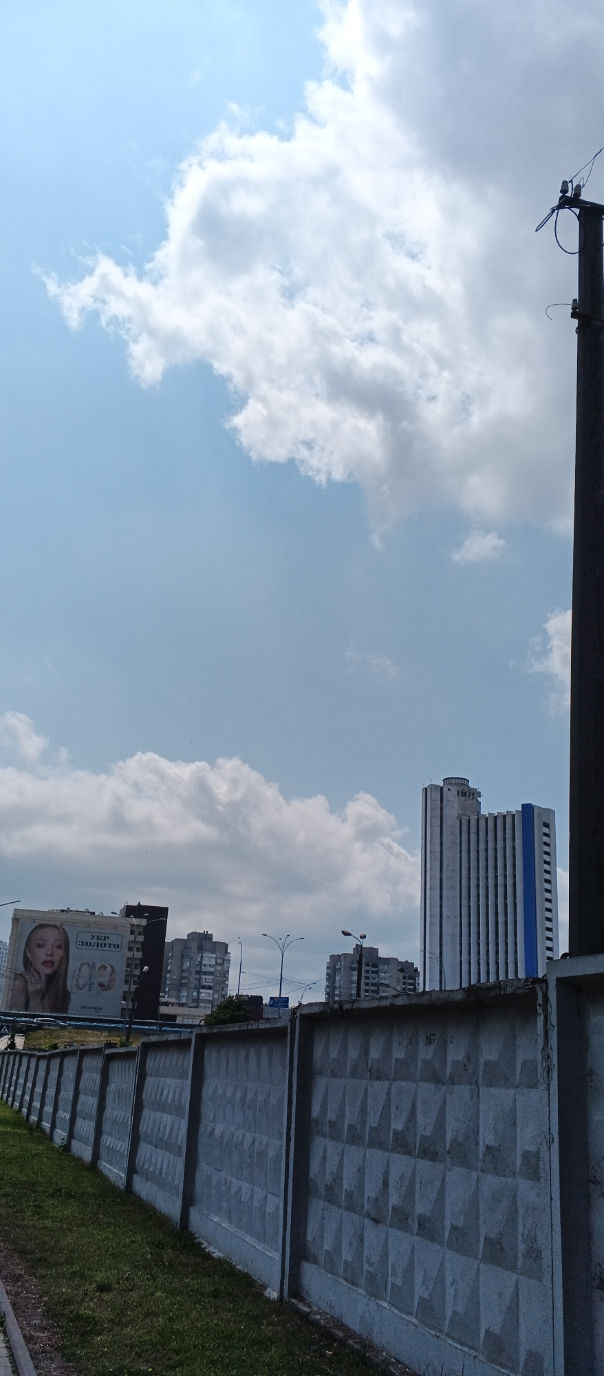The image depicts a street scene in Kyiv, Ukraine, under a partly cloudy sky. In the foreground, a textured concrete fence runs along a grassy area. Beyond the fence, several buildings are visible, including a large billboard featuring a woman and text in Ukrainian. Further in the background, a tall, modern high-rise building with vertical blue accents stands prominently. A utility pole with electrical equipment is positioned on the right side of the frame. The overall impression is of an urban environment on a bright day.