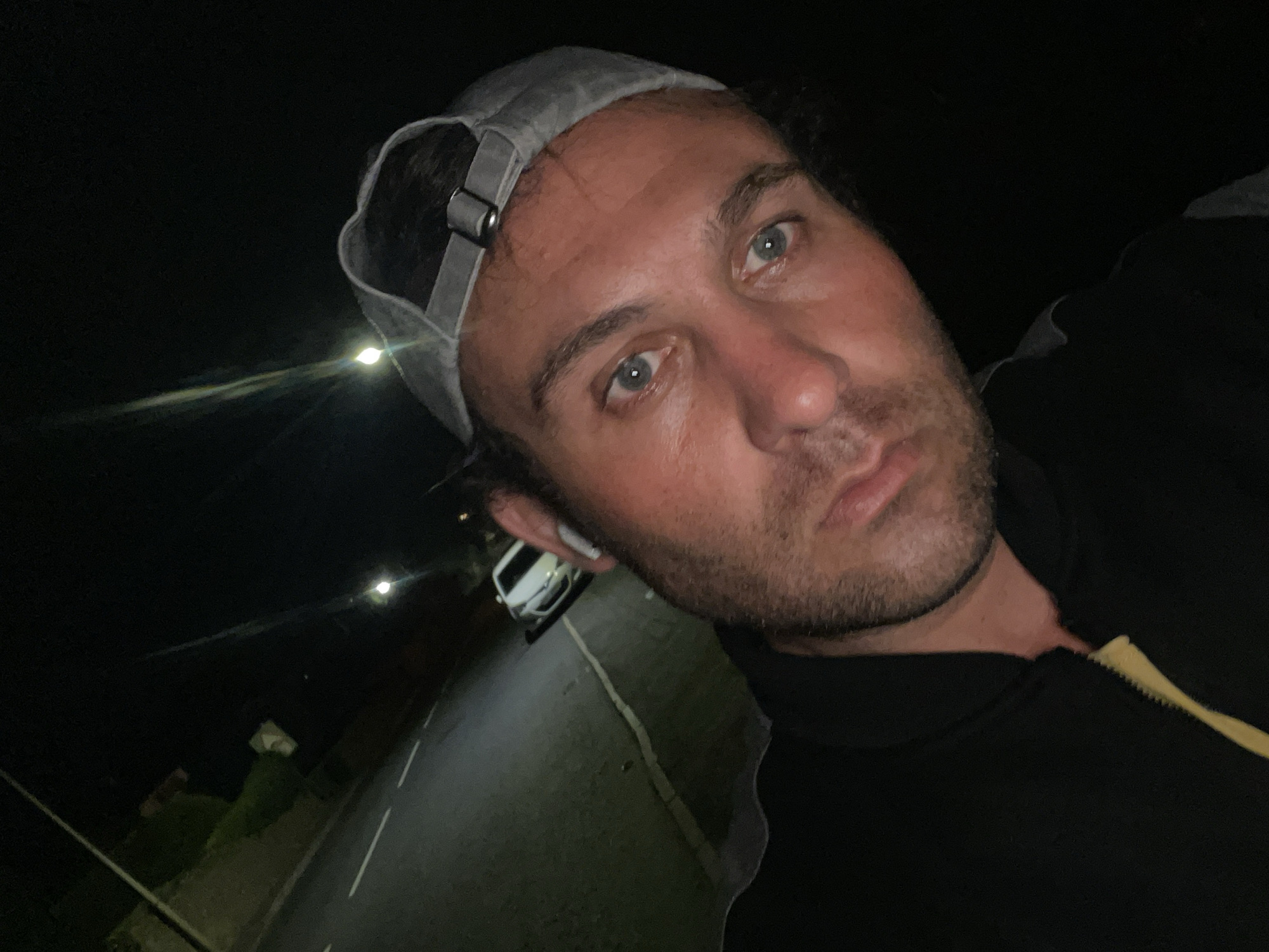 A low-angle, night-time selfie shows a man wearing a grey baseball cap and a black zip-up jacket with a yellow inner lining. He has light blue eyes and stubble. In the background, a street is visible with a white car parked on the side and a single white line marking the road. Streetlights illuminate the scene. The location appears to be a quiet residential street in the city of Armagh, United Kingdom.
