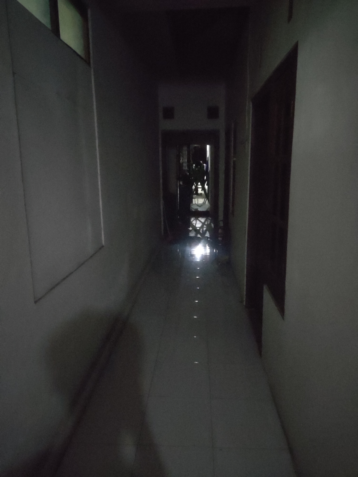 A dimly lit interior hallway in what appears to be a residential building or dormitory in Trihanggo, Indonesia. The hallway is narrow with tiled flooring that reflects a bright light source from the far end. The walls are painted a light color, and there are doors visible on the right side. At the end of the hallway, silhouetted figures can be seen, along with what looks like furniture and possibly a light fixture. The overall impression is one of a dimly lit, possibly abandoned or sparsely populated area.