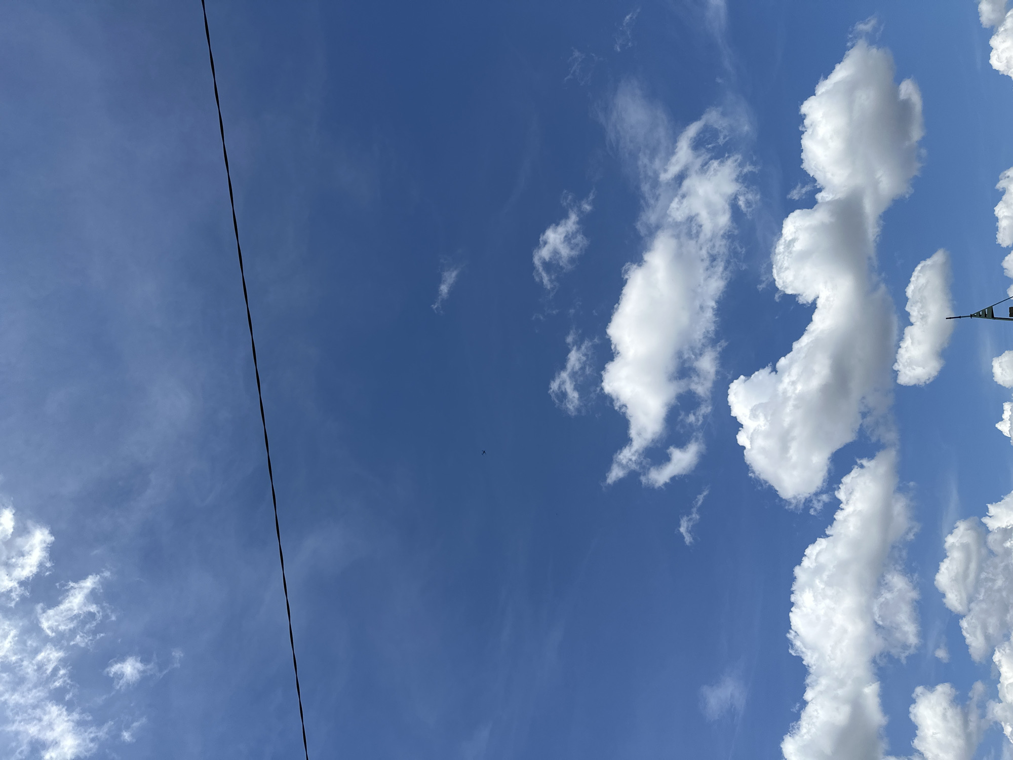 A clear blue sky with scattered white clouds is visible. A dark, thin wire stretches horizontally across the upper portion of the image, dividing the sky. In the distance, a small airplane is silhouetted against the sky. The lower portion of the image shows a glimpse of a structure with a green and white tip, possibly a flag or a marker, suggesting an outdoor setting, potentially in a city like Sofia, Bulgaria, where such structures might be found. The overall impression is a bright, daytime scene with a focus on the sky and atmospheric elements.