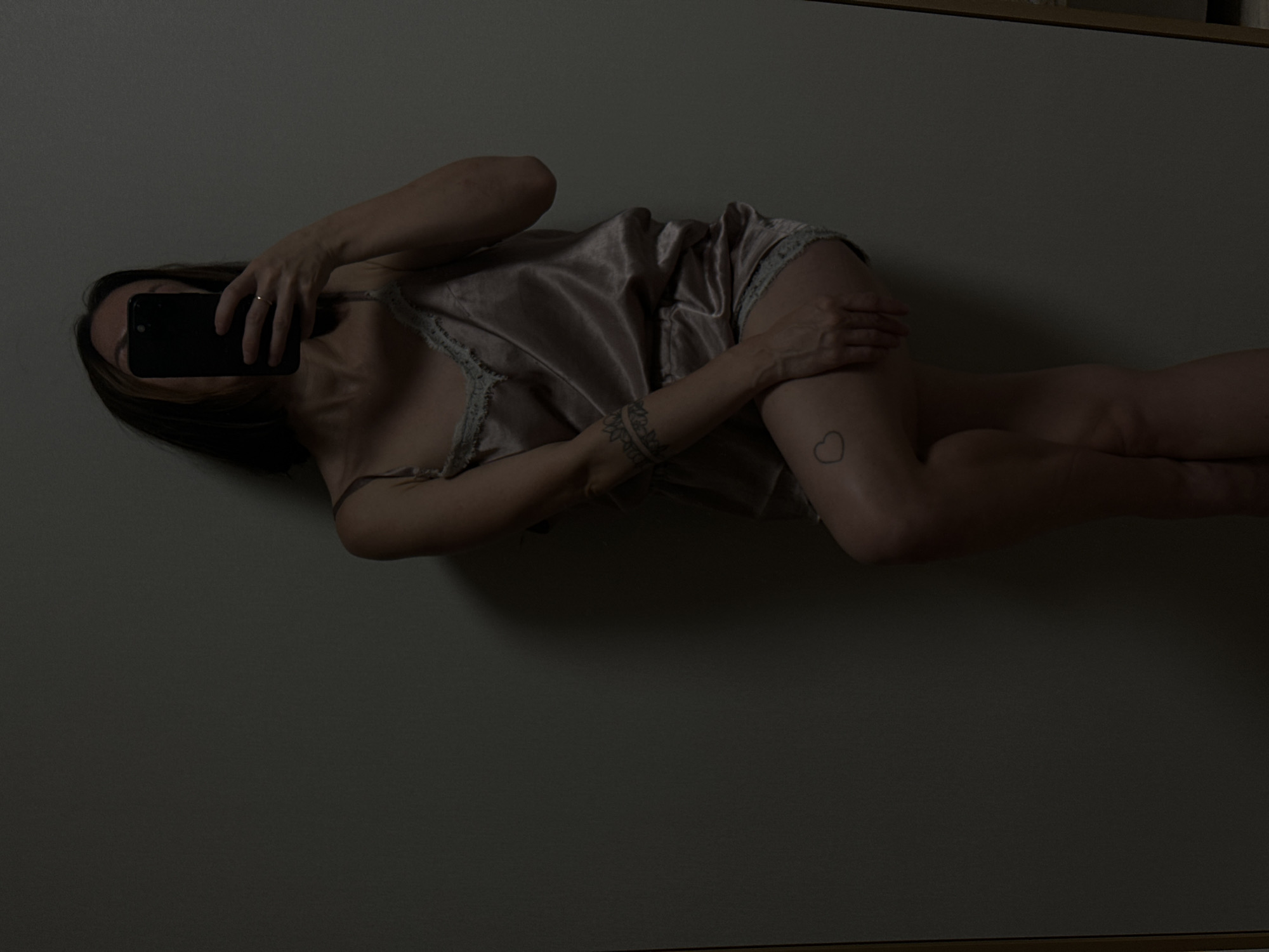 In Votkinsk, Russia, a woman is captured in a dimly lit indoor setting, possibly a bedroom or dressing room. She is standing, leaning against a plain, grey wall, with her left leg bent and her right leg extended slightly forward. She is wearing a light-colored satin camisole and matching shorts, both adorned with delicate lace trim. The camisole features thin spaghetti straps. The woman is holding a dark-colored smartphone in her right hand, angled to take a mirror selfie, obscuring her face. A ring is visible on her right ring finger. Her left arm rests on her bent leg, and a tattoo is visible on her upper left arm and a small heart tattoo is on her left thigh. The lighting is low, casting shadows and highlighting the textures of her clothing and skin. The overall atmosphere is intimate and subdued.