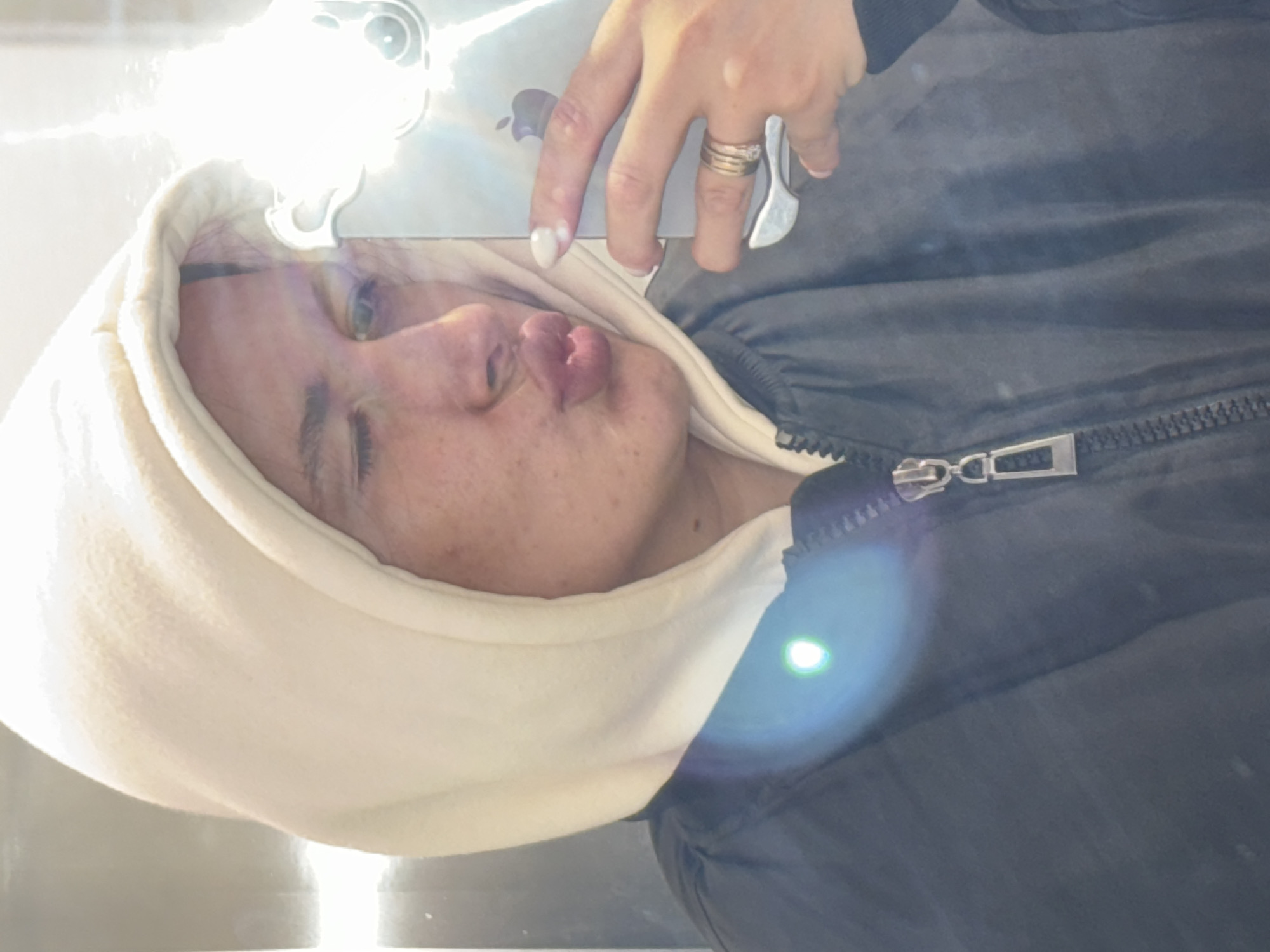 The image, rotated 90 degrees clockwise, shows a person, likely a woman, taking a selfie. She is wearing a light cream or off-white hooded sweatshirt with the hood up, layered under a dark (possibly black or dark grey) zippered jacket. Her face is turned towards the camera, and she is making a "duck face" with puckered lips, conveying a playful expression. Her eyes are partially visible, looking towards the viewer, and there are visible freckles on her skin.
Her right hand is positioned above her head, holding a metallic object, likely a smartphone or tablet, which exhibits a reflective surface. A faint outline of what appears to be an Apple logo is discernible on this device in the cropped hints. Her fingernails are manicured with white tips, and she wears a ring on her ring finger.
The scene is characterized by extremely bright lighting, with significant glare and lens flare originating from the upper left quadrant, suggesting a strong light source directly impacting the camera. This intense light obscures much of the background, making the specific location or setting unidentifiable. The activity depicted is a casual, self-portrait photograph.