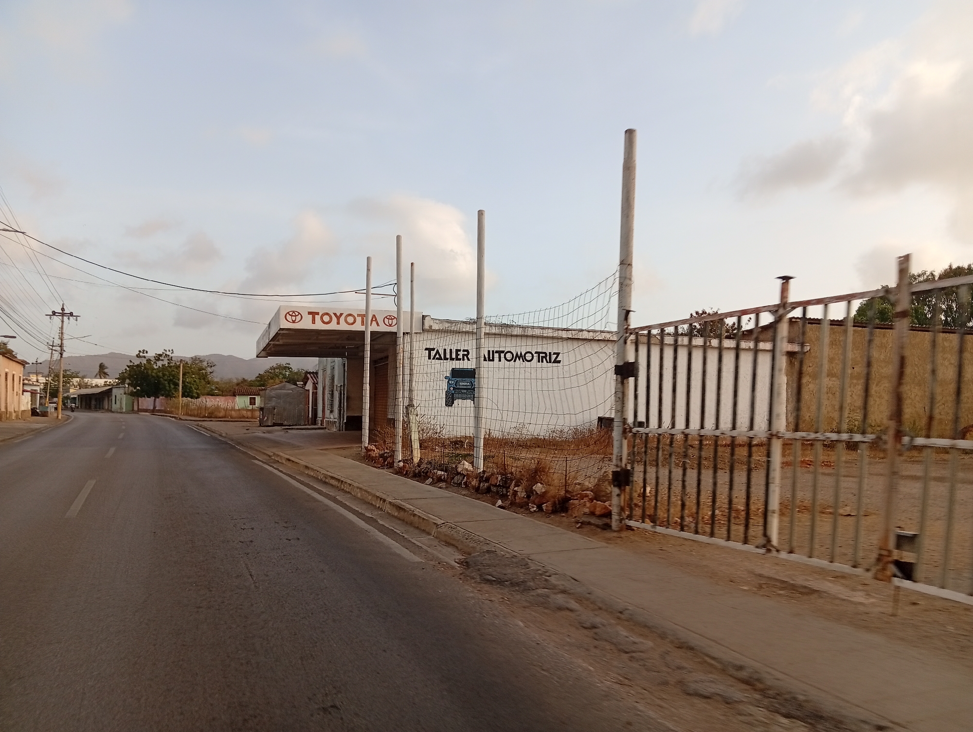 The image depicts a street scene in Juan Griego, Venezuela. A Toyota dealership or service center ("Taller Automotriz") is visible on the right side of the road. A partially dilapidated fence runs along the perimeter of a property, partially obscuring the view. The road appears wet or recently cleaned. Power lines run overhead, and various buildings and vegetation are visible in the background. The sky is overcast with light clouds.