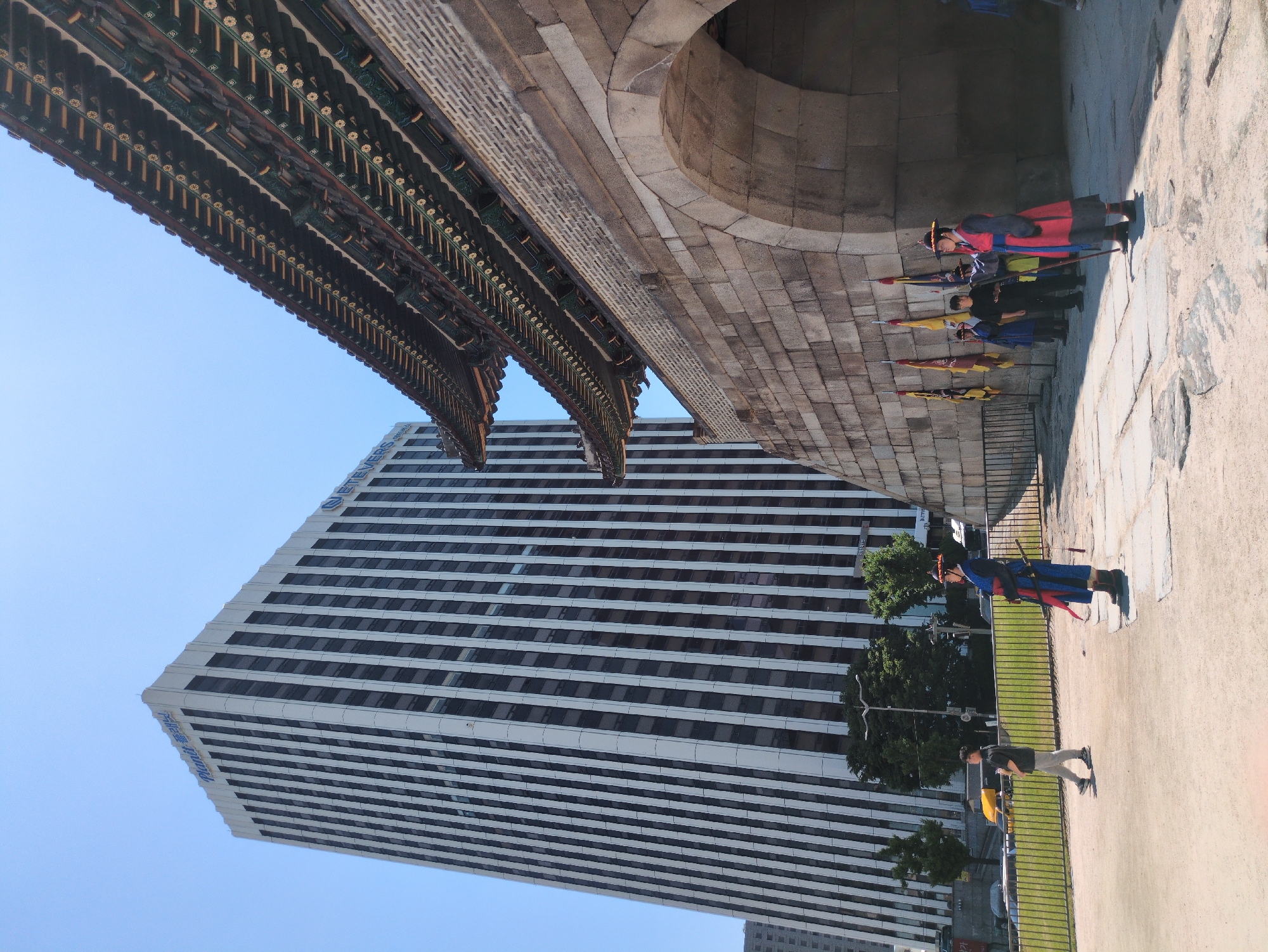 The image is taken at a low angle in Seoul, South Korea, showing a blend of traditional and modern elements. The architecture of the Sungnyemun Gate is visible, featuring a stone archway and a highly decorated traditional roof. Within the archway are guards in traditional Korean attire, holding spears and flags. A modern, striped high-rise building is visible in the background. Some modern pedestrians are present on the right-hand side of the image. The sky is clear and blue.