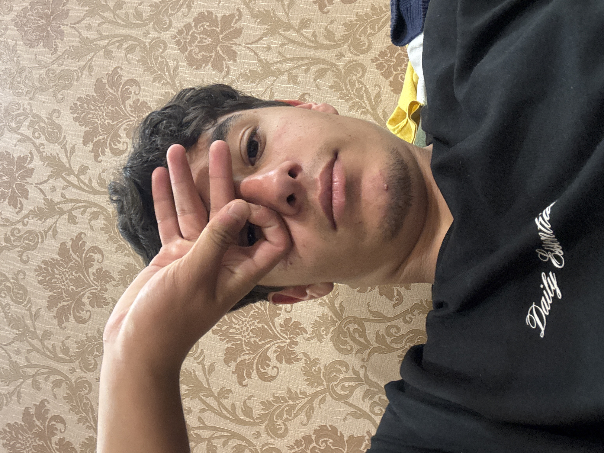A man is posing indoors, likely in a room with wallpaper featuring a floral pattern. He is wearing a black top and has his hand near his face, making a gesture. The specific location in Yerevan, Armenia cannot be determined based on the image.