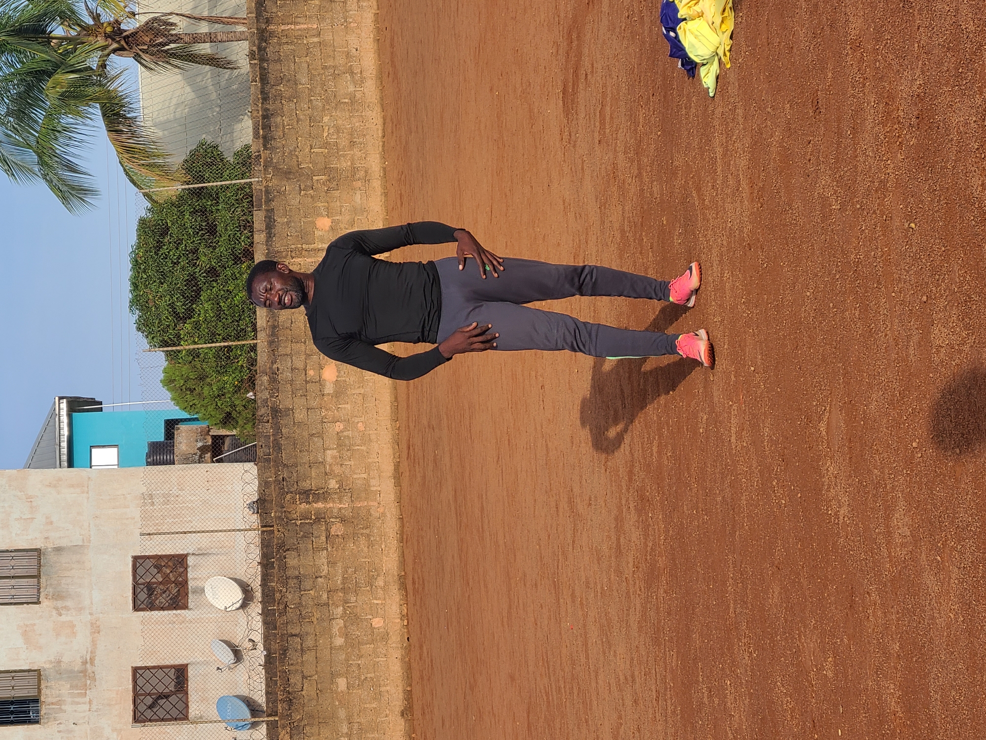 In Barakin Lerawi, Nigeria, a Black man stands on a reddish-brown dirt field, likely a sports ground, under a clear sky. He is wearing a long-sleeved black athletic shirt and dark gray sweatpants, with bright pink athletic shoes. His left hand rests on his hip, and his right hand is bent at the elbow, with his fingers positioned near his chest. He appears to be looking off to his right, with his mouth slightly open. Behind him, a low brick wall runs horizontally across the frame. Beyond the wall, a chain-link fence is visible, separating the field from a more urbanized area. In the background, there are buildings, including a light-colored structure with multiple windows, a bright blue building, and a building with a metallic roof. Vegetation, including trees and a palm tree, is also present. To the right of the man, a pile of folded yellow and blue athletic apparel lies on the ground. The man casts a shadow on the dirt field.