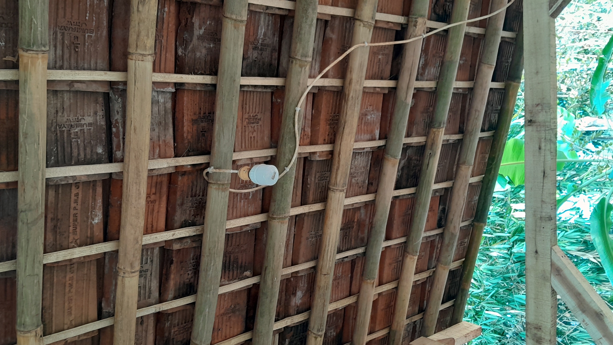 This is a close-up, low-angle, vertical, full-frame shot of a rustic roof structure, likely located in Sumedang, Indonesia. The roof is constructed with terracotta tiles, supported by a framework of bamboo poles and thinner wooden slats. The bamboo poles are arranged horizontally, while the wooden slats are positioned perpendicular to them, creating a grid-like pattern. Some of the terracotta tiles are visible beneath the bamboo and wood, showing signs of age and wear. A single light bulb, with a light blue shade, is suspended from the bamboo structure by a cord. The cord runs diagonally across the frame, secured by small white clips attached to the bamboo. In the background, out of focus, are lush green foliage, suggesting an outdoor setting surrounded by vegetation. The lighting is dim, indicating it might be overcast or late in the day. There are no people or animals visible in the image. The scene appears to be a part of a simple, possibly traditional, building.