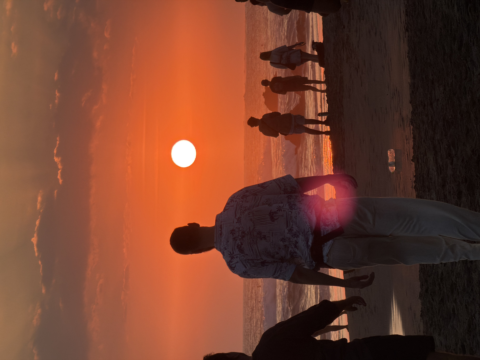 At sunset on the beach in North Kuta, Indonesia, a group of people are silhouetted against a fiery orange sky. The sun, a bright white disc, is low on the horizon, casting a warm glow over the scene. Waves gently lap at the shore, and a few rocks emerge from the water. In the foreground, a man stands with his back to the camera, wearing a patterned shirt and light-colored pants. To his left, another person is partially visible, wearing dark clothing. Further down the beach, a small cluster of individuals watches the sunset, their forms indistinct against the vibrant backdrop. A plastic bottle lies on the wet sand near the water's edge. The overall atmosphere is one of peaceful observation and natural beauty.