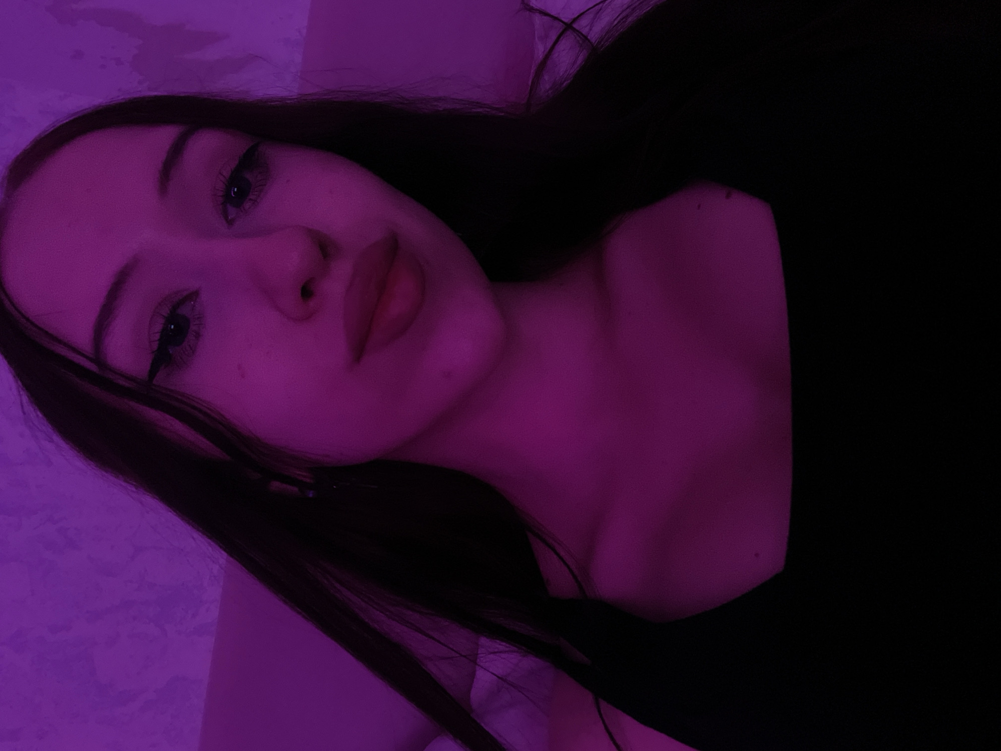 A young woman with long dark hair and winged eyeliner is shown in a close-up selfie. She is wearing a black top and is illuminated by purple lighting. The background appears to be a textured wall. The image does not contain any information that can link it to the city of Votkinsk, Russia.